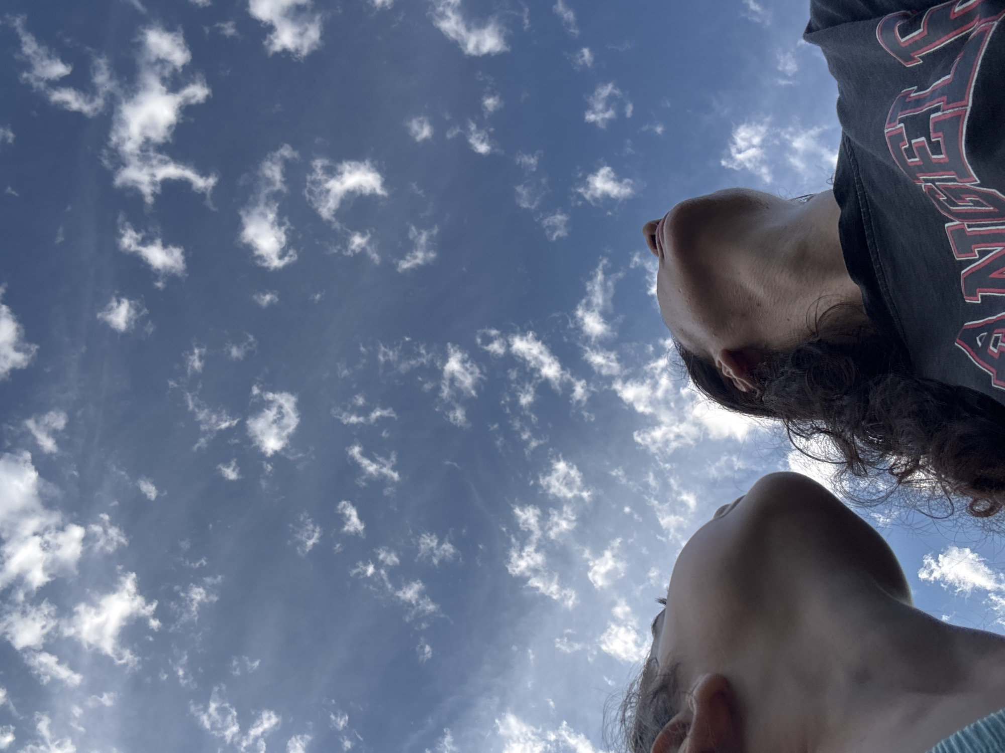 Two people are looking up at the sky. The person on the left is viewed from the side, and their face is partially obscured by their head. They have dark hair. The person on the right is facing more towards the camera, with their head tilted back and looking upwards. They have dark, curly hair and are wearing a dark t-shirt with red and white lettering. The sky is a bright blue with numerous scattered white clouds. The location is not identifiable as Sochi, Russia from the image provided.