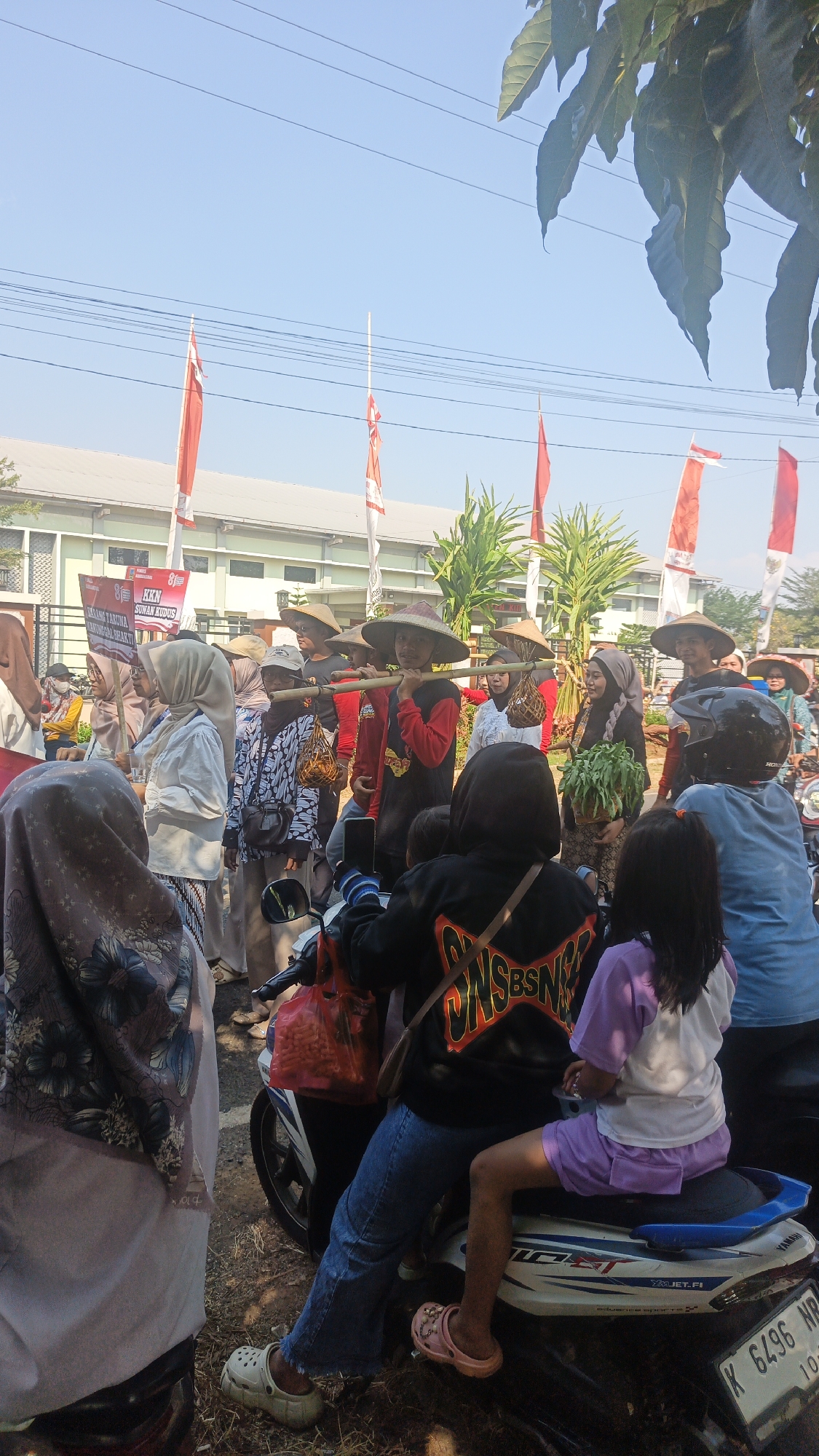 The image shows a group of people standing outside a building, some of them holding bamboo poles with baskets attached. Many of the people are wearing hats and traditional clothing, and some are carrying plants or bags. There are red and white flags in the background. The scene appears to be a community event or festival.