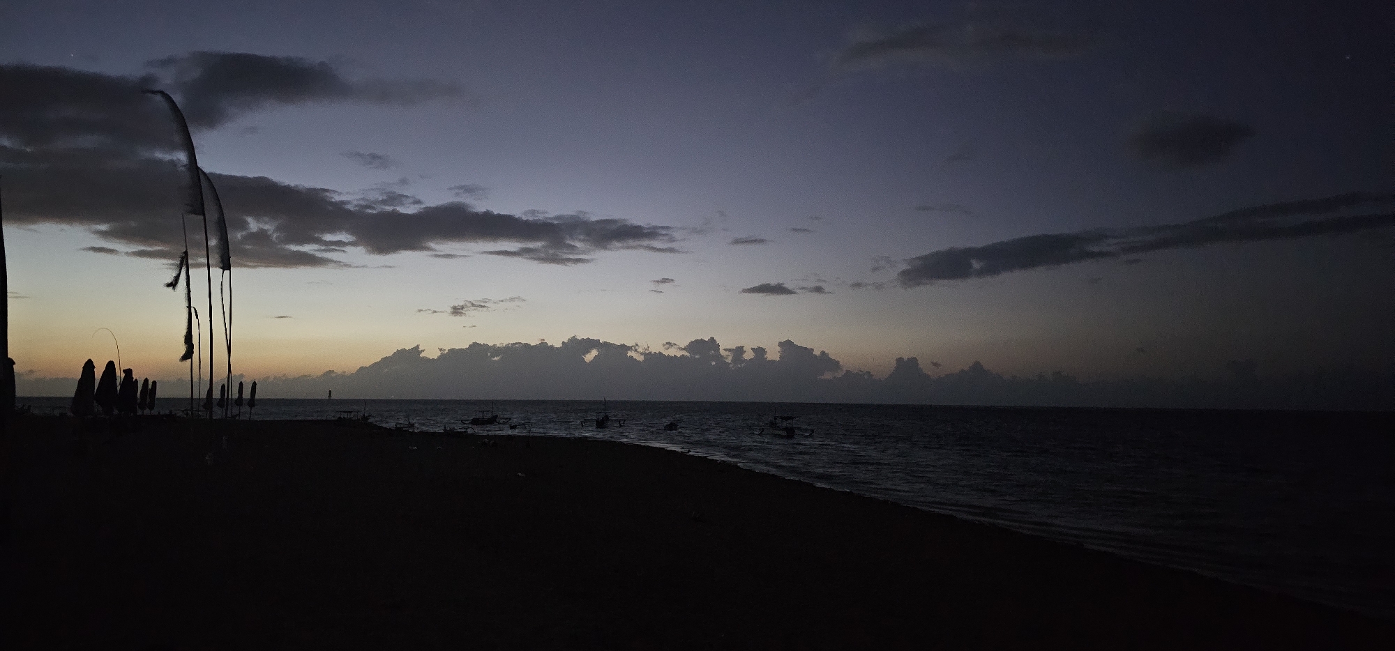 The image shows a beach scene in Denpasar, Indonesia, at dusk or dawn. The sky is a mix of dark blue and lighter orange hues, with clouds scattered across the horizon. The beach is dark, with silhouettes of flags and beach umbrellas on the left side. The ocean is visible in the middle ground, with several small boats floating on the water. The overall scene is dim and tranquil.