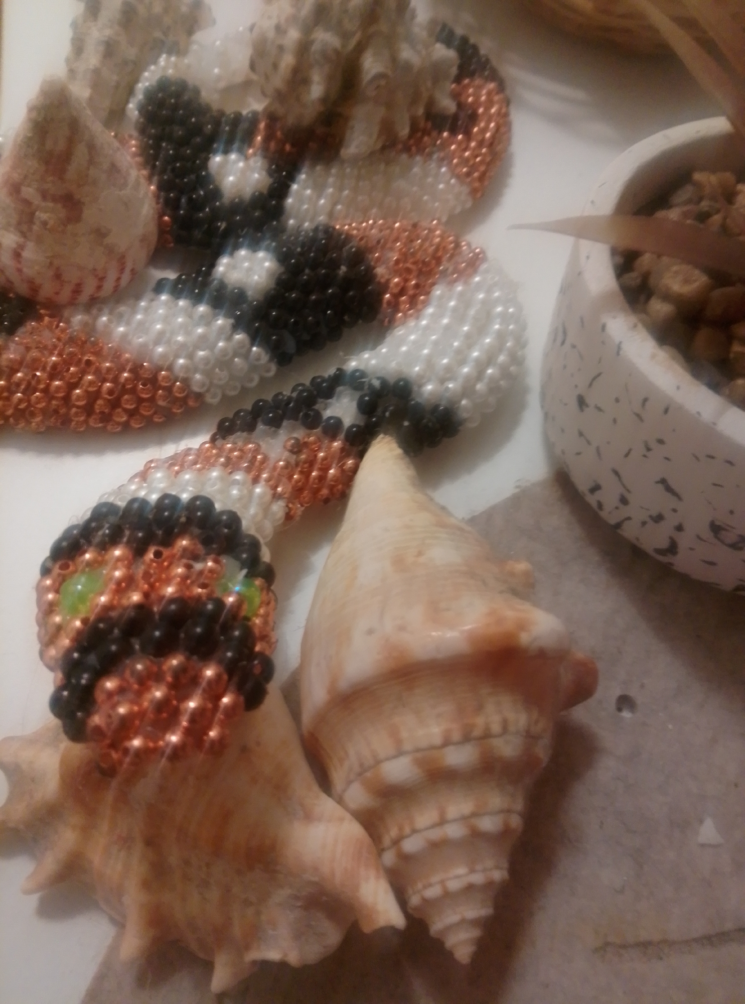 The image shows a close-up, top-down view of a collection of seashells and a beaded snake on a white surface. The beaded snake is made of black, white, and copper-colored beads and appears to be coiled around some of the seashells. Two large, spiral-shaped seashells are prominent in the foreground. In the background, there is a white pot with dark speckles containing small stones and a plant with long, thin leaves. The lighting suggests it's indoors. There are no people present in the image. The location is specified as the city of Wedel, Germany.