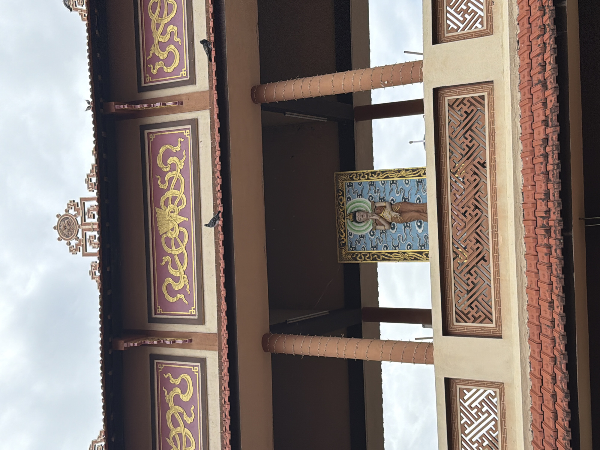 The image shows a low-angle, eye-level view of a building with distinct architectural features, likely a temple or cultural site in Hoi An, Vietnam. The sky above is overcast with a pale, cloudy texture. The building's facade is adorned with decorative elements, including what appear to be elaborate golden dragon motifs against a deep purple background, set within rectangular panels. Above these panels, a tiered, ornate structure with symbolic carvings is visible.
The building has a prominent, tiled roof in a reddish-orange hue, characteristic of traditional Vietnamese architecture. Below the upper facade, a covered walkway or veranda is supported by several round, reddish-brown columns that are wrapped with a lighter-colored rope or string in a spiraling pattern.
In the center of the image, within this covered area, a framed artwork or statue is displayed. It depicts a figure with arms raised, possibly in a prayer or blessing gesture, set against a backdrop of blue clouds. The figure appears to be wearing robes and has a serene expression.
Below the artwork, there is a decorative latticework panel made of brick or stone, featuring intricate geometric patterns. This panel is framed by a lighter colored border.
Two pigeons are visible on the roofline of the upper facade, and another pigeon is perched on the ledge below the decorative panels. The overall impression is one of a culturally significant and well-maintained structure.