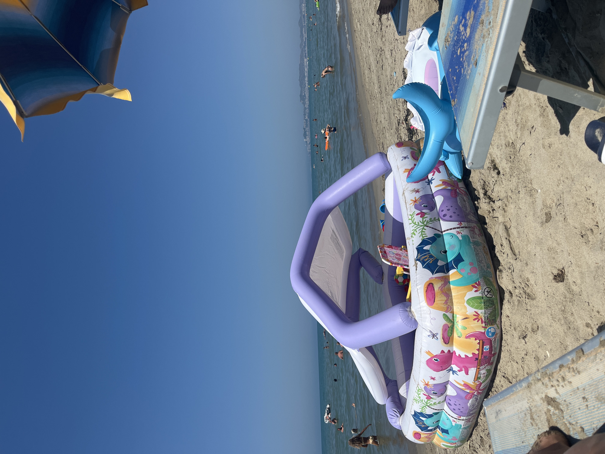 The image depicts a sunny day at a beach in Bashkia Durrës, Albania. In the foreground, a child's inflatable pool with a dinosaur design is placed on the sand. A blue inflatable toy is next to the pool, and a beach chair is visible in the lower right corner, partially covered in sand. In the background, the sea is calm with several people swimming and wading in the water. The horizon shows a faint outline of buildings, suggesting a coastal city. The sky is clear and blue.