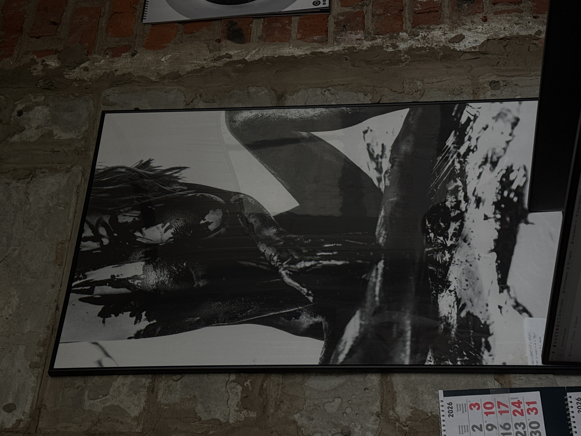 A black and white framed photograph hangs on a weathered brick wall in what could be a space in or near the city of Chaikovsky, Russia. The image appears to depict a partially obscured human figure covered in what resembles mud or dark paint. Part of a calendar displaying the year 2026 is visible below the photograph. A corner of a computer monitor also enters the frame on the right side of the photograph. The location is an interior with an aging, possibly industrial, aesthetic, judging by the texture of the brick and plaster walls.