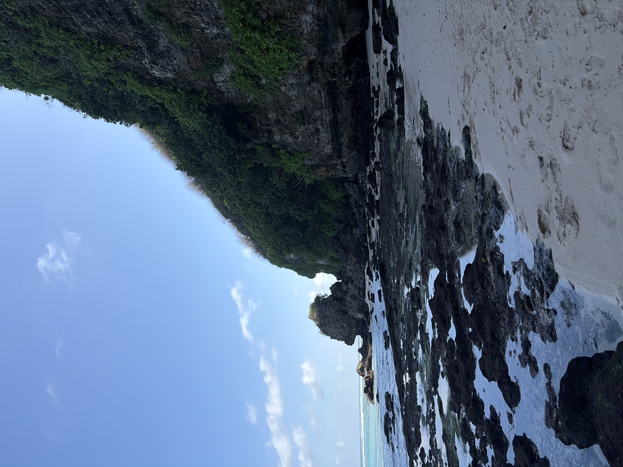 An outdoor, eye-level, medium-shot photograph captures a rocky shoreline and cliff face on a clear day in Kuta Selatan, Indonesia. The turquoise ocean water gently laps against numerous dark, moss-covered rocks that are partially submerged and scattered across the foreground. A sandy beach with visible footprints stretches along the right side of the frame. To the left, a distinctive, large rock formation rises from the sea. Dominating the background is a steep, rugged cliff face covered in dense green vegetation. The sky above is a bright, cloud-dappled blue.
