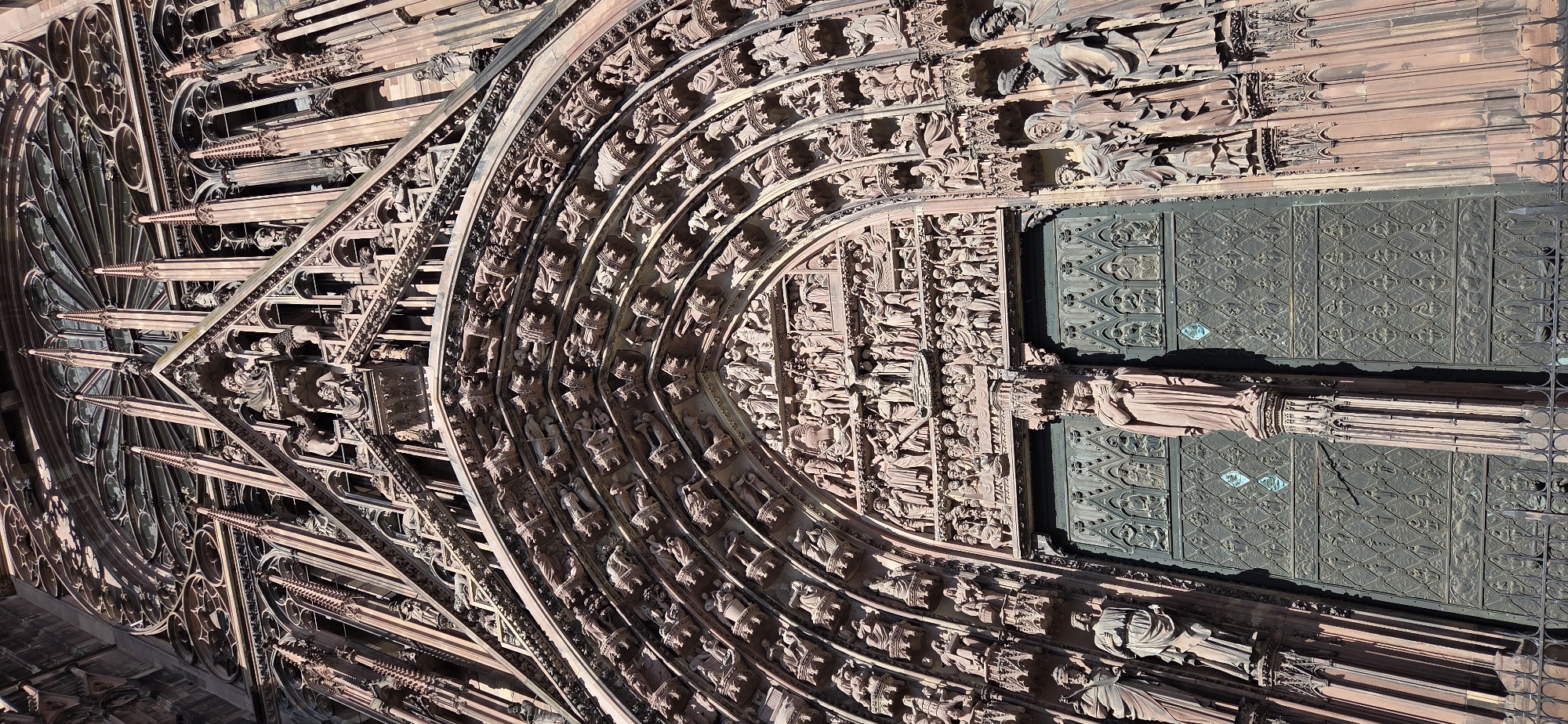 The image depicts a close-up, low-angle view of the ornate facade of Strasbourg Cathedral, officially known as the Cathédrale Notre-Dame de Strasbourg, located in Strasbourg, France. The architecture is predominantly Gothic, characterized by intricate stone carvings, arched doorways, and towering spires. The facade is adorned with numerous sculpted figures, depicting religious scenes and individuals, forming multiple layers of detail. The main entrance features large, metal doors with relief sculptures. The material of the cathedral appears to be a reddish sandstone, which gives it a warm hue, particularly where the sunlight strikes it. The overall scene conveys a sense of grandeur and historical significance.