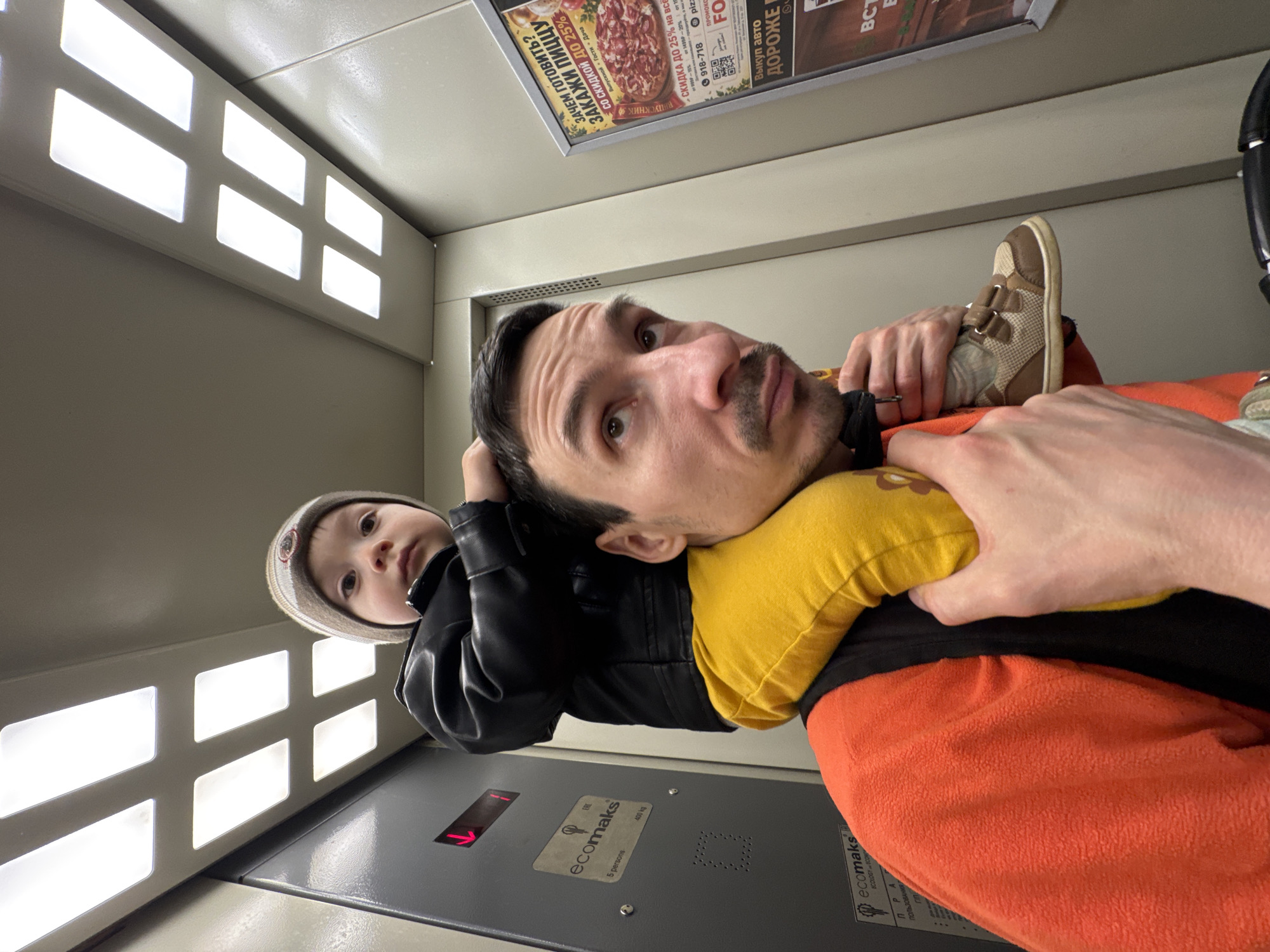 A man is in an elevator holding a young child on his shoulder. The man is wearing an orange shirt and has a mustache. The child is wearing a jacket and hat. An advertisement for pizza and auto sales is visible at the top. The elevator appears to be in Izhevsk, Russia, based on the advertisement. The elevator has a sign that reads "ecomaks 5 persons 400 kg."