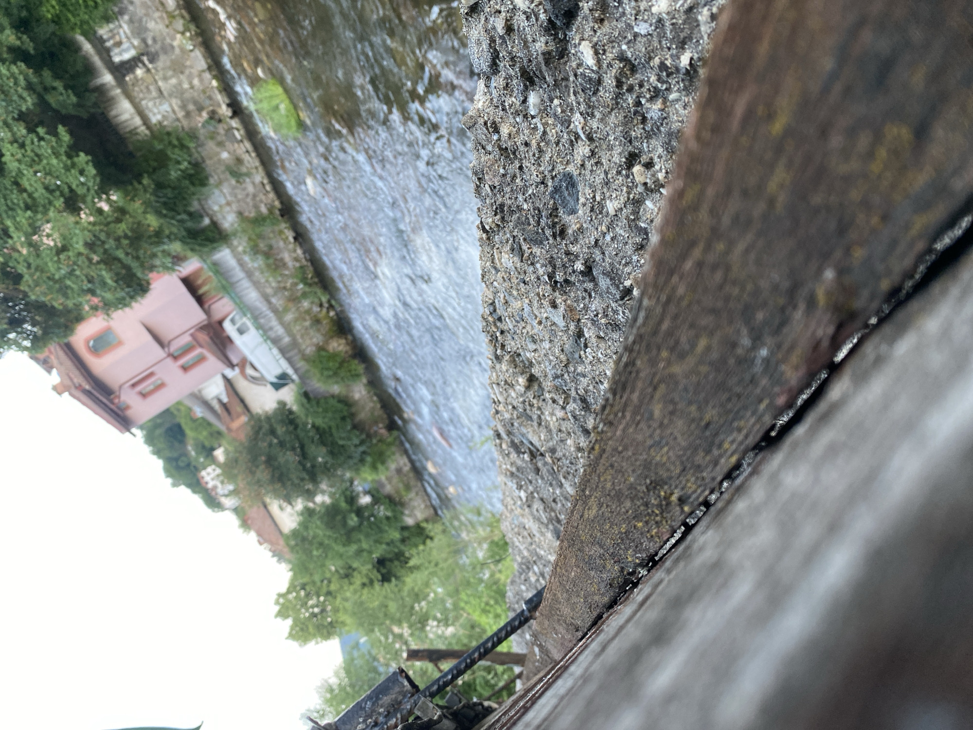 The image shows a narrow, rocky river flowing through a town. Buildings with red-tiled roofs and pink facades are visible on the hillside overlooking the river, with lush green trees interspersed among them. In the foreground, there is a rough, concrete embankment with a wooden railing that appears weathered and aged. The scene is captured from a low angle, looking across the river towards the town. The water is choppy, suggesting a current. The sky is overcast and bright. This scene depicts a riverside area in Smolyan, Bulgaria, likely a part of the town where the river runs through the urban landscape.