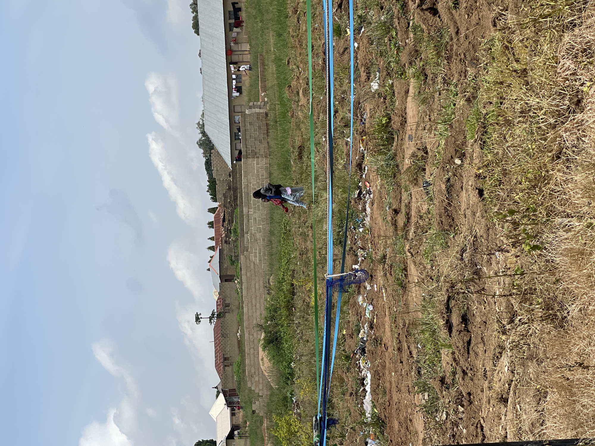 This media file depicts an outdoor scene in Durumi, Nigeria, likely during daylight hours under a cloudy sky. The setting appears to be a somewhat unkempt area with buildings constructed from brick and metal roofing visible in the background, some with laundry hanging outside. In the foreground, there is a patch of disturbed earth with dry, overgrown grass and scattered debris, including plastic waste. Several blue and green ropes or pipes are strung across the scene, possibly indicating some form of construction or utility work.
In the mid-ground, two people are walking together. One is wearing a dark jacket over a red shirt and light-colored pants, carrying a dark bag on their shoulder. The other person is wearing a light-colored top and dark pants. Their posture and the way they are walking suggest they are engaged in a casual movement. The overall impression of the scene is one of everyday life in a developing or rural area, with a mix of residential buildings, undeveloped land, and visible infrastructure. There is no visible text in the image.