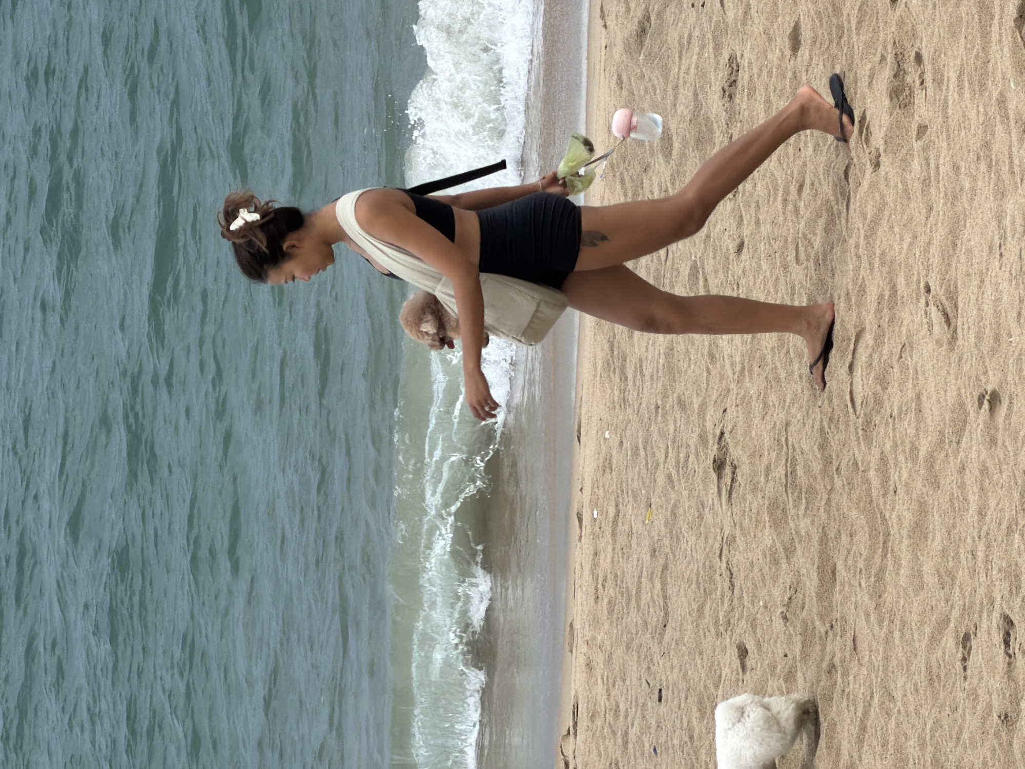 A woman with dark hair tied up in a bun with a white clip walks along the sandy shore of a beach in Nha Trang, Vietnam. She is wearing a black sports bra and black shorts. A light-colored sling bag is slung over her right shoulder, carrying a small, light brown dog. In her right hand, she holds a green poop bag dispenser and a water bottle. A white dog is visible in the background to the left. The ocean with moderate waves is to the left of the woman, with the sandy beach occupying the foreground and midground. The weather appears overcast.