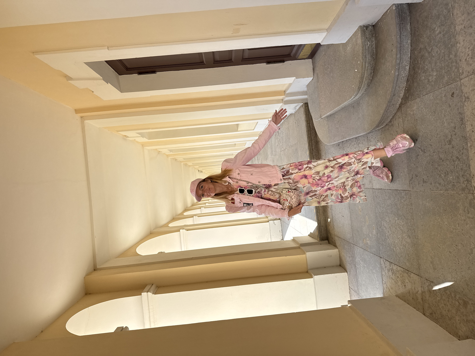 The image shows a woman standing in an arched corridor with yellow walls. She is wearing a pink denim jacket, a floral dress, and pink sneakers. She is also carrying a small purse and wearing a pink baseball cap. The corridor has a stone floor and a dark wooden door on the right side of the frame. The woman is smiling and extending her hand towards the viewer. The architecture of the corridor suggests a historical building, possibly a palace or a museum. Based on the architectural style, the location is likely the Grand Menshikov Palace in Lomonosov, Russia, known for its classical design and extensive colonnades.