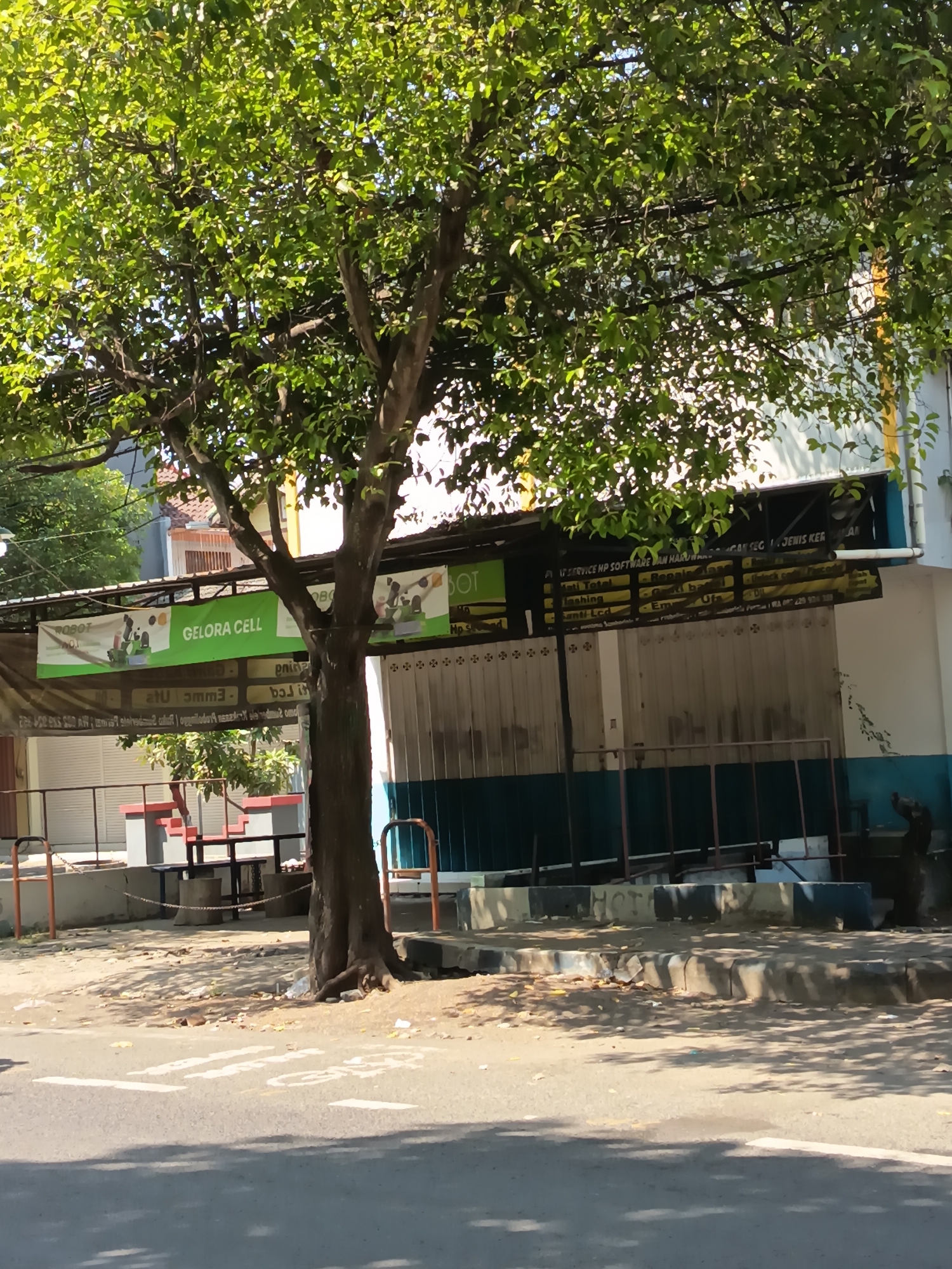 A medium shot captures a sun-drenched street scene in front of a row of shops in Kandangjati Kulon, Indonesia. A large tree with dense green foliage stands prominently in the foreground, its trunk partially obscuring the view of the shops. The shops have metal roll-up doors, some of which are open to reveal the interiors. Above the doors, banners and signs are visible, one of which reads "GELORA CELL". The pavement in front of the shops is shaded by the tree, with scattered debris. A sidewalk with a raised curb separates the shops from the street, which shows faded white markings. Orange metal barriers are positioned along the sidewalk. The environment appears to be an urban or semi-urban area, likely during daylight hours.