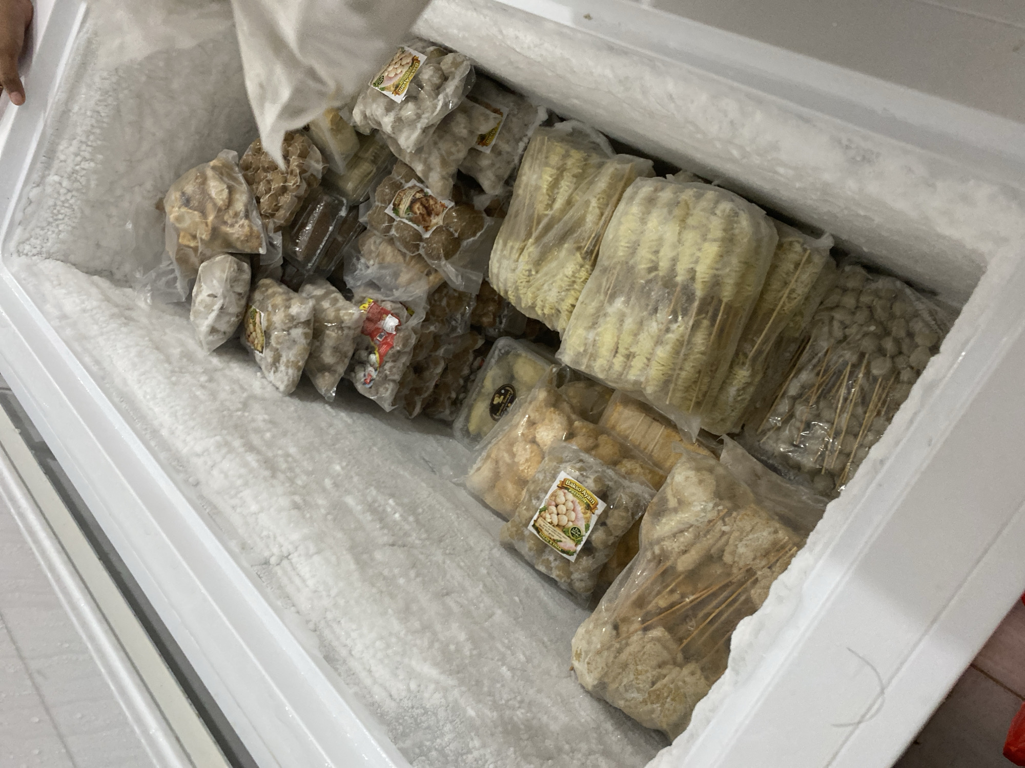 The image shows the interior of a chest freezer filled with various frozen food items, primarily packaged and frozen in plastic. The freezer compartment is heavily frosted, indicating its cold temperature. The food items are arranged in stacks and piles, with many packaged in clear plastic bags, some with visible labels. Among the visible items are what appear to be meatballs, skewers of battered and fried items, and possibly other processed meat or seafood products. A hand is partially visible in the upper right corner of the image. The scene suggests a food storage or preparation environment. Without further context or visible landmarks, the specific location in the city of Payatieng, Indonesia, cannot be determined from the image alone.