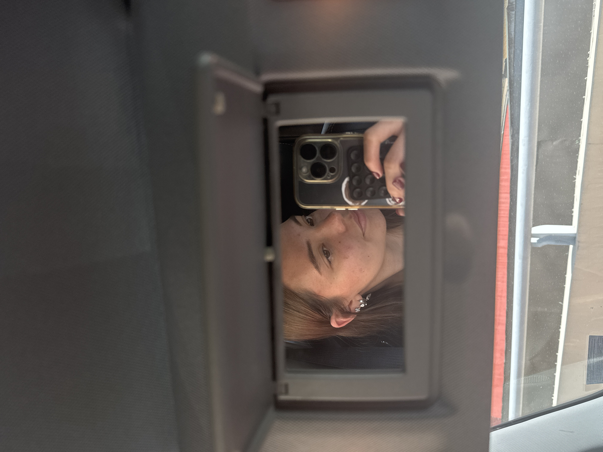 A person with light skin, freckles, and light brown hair reflected in a car's vanity mirror holds a cell phone. The phone has a black case with small raised circles on the back and three camera lenses. The mirror is attached to a gray car sun visor. A blurry view of a vehicle and a road is visible through the car window, suggesting an urban location. The city of Puebla, Mexico, is not definitively identifiable from the provided image.