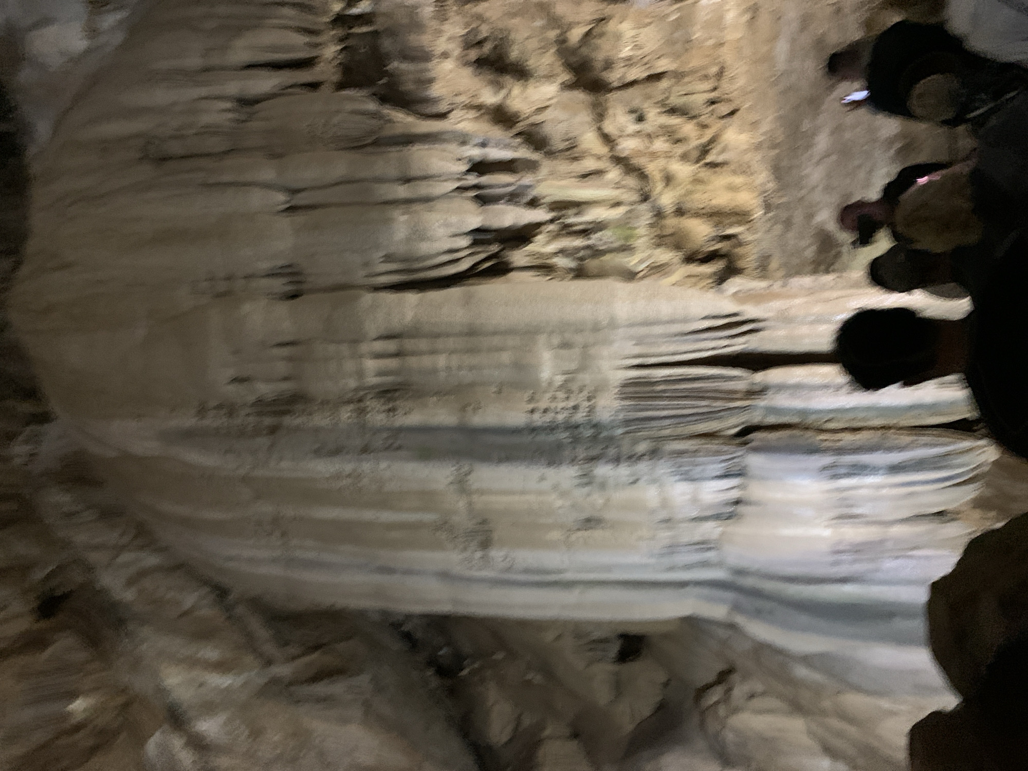 The image shows a poorly lit interior of a cave, likely in Inkiti, Abkhazia. Layered rock formations, possibly stalactites or stalagmites, dominate the scene. Several blurred figures are partially visible on the right side of the frame. They appear to be tourists or explorers. The overall environment suggests a natural cave system with geological features.