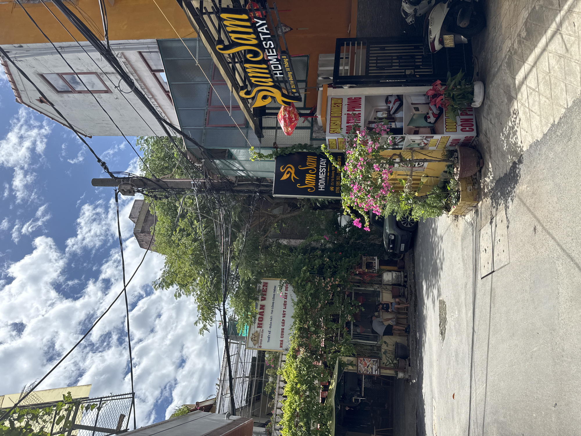The image captures a street scene in Hoi An, Vietnam, on a partly cloudy day. In the foreground, a paved street with a manhole cover is visible, casting dappled shadows. To the right, a white and silver scooter is parked next to a dark metal gate. A yellow and orange building with various signs is prominent. One sign, illuminated by a red lantern, reads "Sam Sam HOMESTAY" in yellow lettering. Below this, a poster displays images of hotel rooms and text that includes "ROOM AVAILABLE" and "CON PHONG." Another black sign with a yellow swan logo and "Sam Sam HOMESTAY" is mounted on a green vine-covered structure to the left.
Further into the scene, a dark colored SUV is parked on the street. A building with a large sign in Vietnamese, "HOÀN VIÊN" (meaning "Complete Happiness" or "Reunion"), is situated behind dense green foliage and flowering plants. This sign advertises "THIẾT KẾ - GIÁM SÁT - THI CÔNG TRÌNH" (Design - Supervision - Construction) and "NHÀ CUNG CẤP VẬT LIỆU XÂY DỰNG" (Building Material Supplier). Two individuals are visible near the entrance of this building, one standing and one seated. Overhead, a tangle of electrical wires crisscrosses the sky, a common sight in Vietnamese cities. The architecture suggests a blend of older structures with newer additions, characteristic of urban areas in Vietnam.