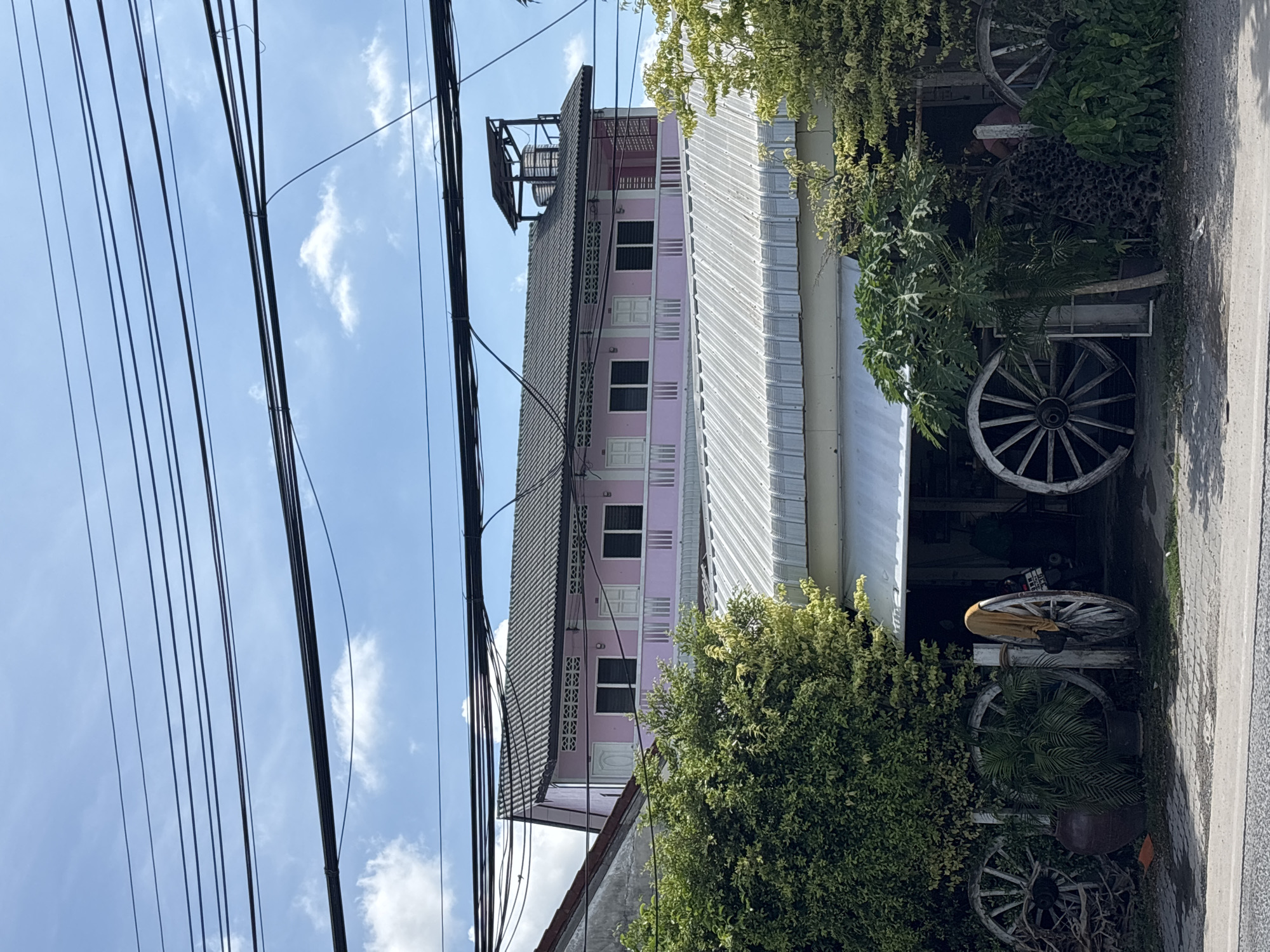 The image depicts a street scene in Choeng Thale, Thailand, showcasing a three-story building painted pink with white trim. The building has a grey metal roof. Atop the building is a metal structure holding a water tank. Utility lines are prominent in the foreground, obscuring portions of the blue sky with scattered clouds. To the right of the building, a covered area exhibits wooden wagon wheels and various plants. The composition is vertical, showing a mix of urban construction and natural elements. The photo appears to be taken during the day.
