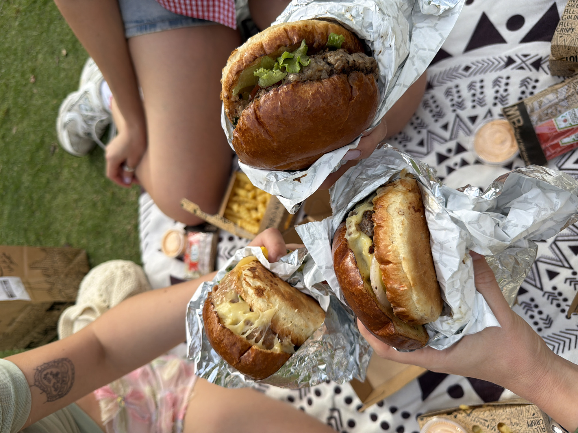 Here is a description of the scene in Tel-Aviv, Israel:
**Scene:** Outdoor picnic on a patterned blanket.
**People:** At least three individuals are present, identifiable by arms and legs holding food.
**Subjects:** Three burgers wrapped in foil; one appears to be cut in half. A container of fries and dipping sauce is also visible.
**Objects:** The blanket is a black and white geometric pattern. There are containers and food packaging with English writing.
**Actions:** The people are holding the burgers as if to eat them.
**Interactions:** The individuals are likely sharing a meal together.
**Environment:** The picnic takes place on grass, suggesting a park setting. The setting is likely Tel Aviv, Israel, based on the English/Hebrew product packaging and the type of food present.