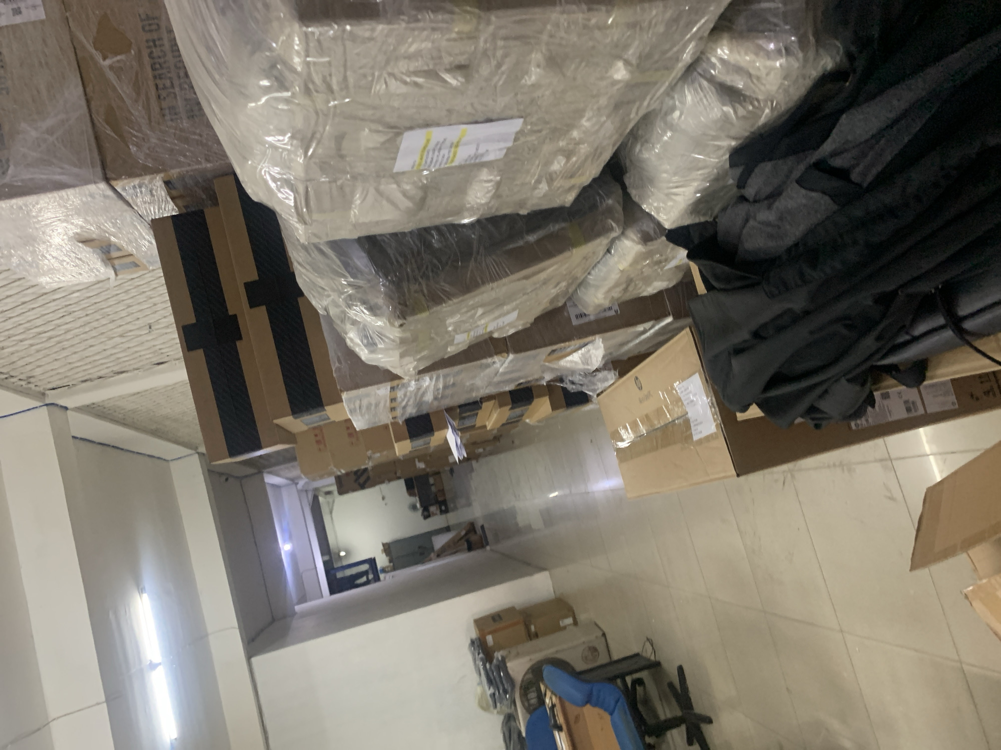 The image shows a storage room filled with stacks of boxes, some wrapped in clear plastic. The room appears to be an office or warehouse space. In the foreground, there are dark-colored garments, possibly clothing, laid out on top of boxes. To the left, a blue office chair is positioned in front of a desk. The walls are white, and fluorescent lighting is visible on the ceiling. The floor is tiled. In the background, more stacks of boxes line the walls and extend down a hallway, which leads to a dimly lit area with what appear to be metal cabinets. The overall impression is one of a cluttered but organized storage area.
Without further information or visual cues specific to Palembang, Indonesia, it is impossible to pinpoint the exact location within the city. The image does not contain any identifiable landmarks, street signs, or cultural elements that would confirm its location in Palembang.
