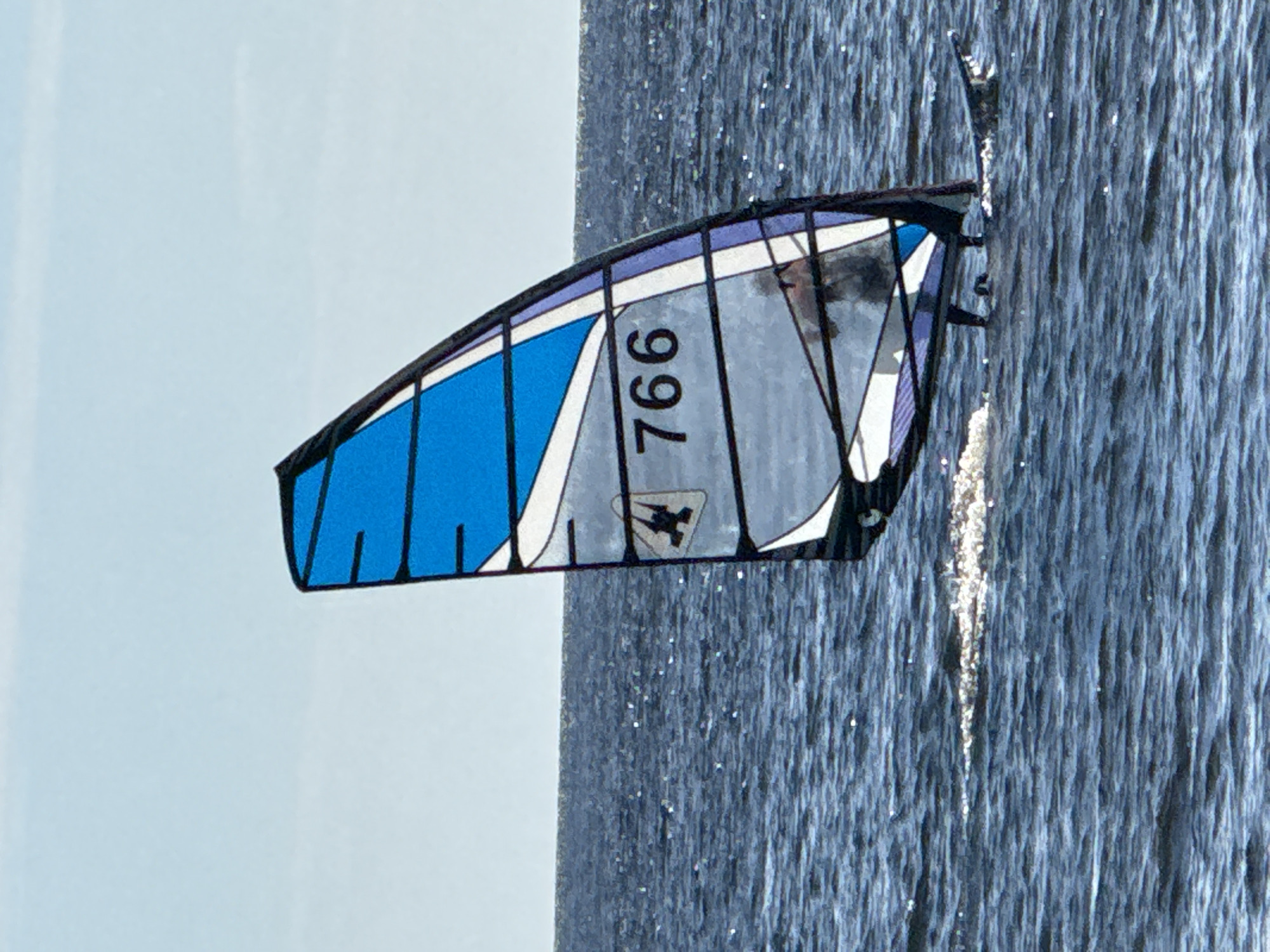 A windsurfer is seen from the side in the middle of a body of water. The windsurfer is a person with dark hair and is wearing a dark wetsuit. They are standing on a board and holding onto a sail, which is blue and white with the number 766 printed on it. The sail is partially transparent, allowing a view of the windsurfer's legs and the board. The water is dark blue and choppy, with white caps visible. The sky is light blue and clear. The scene appears to be taking place outdoors, likely on a sunny day. The image does not provide enough information to determine the specific location within Zelenaya Roshcha, Russia.
