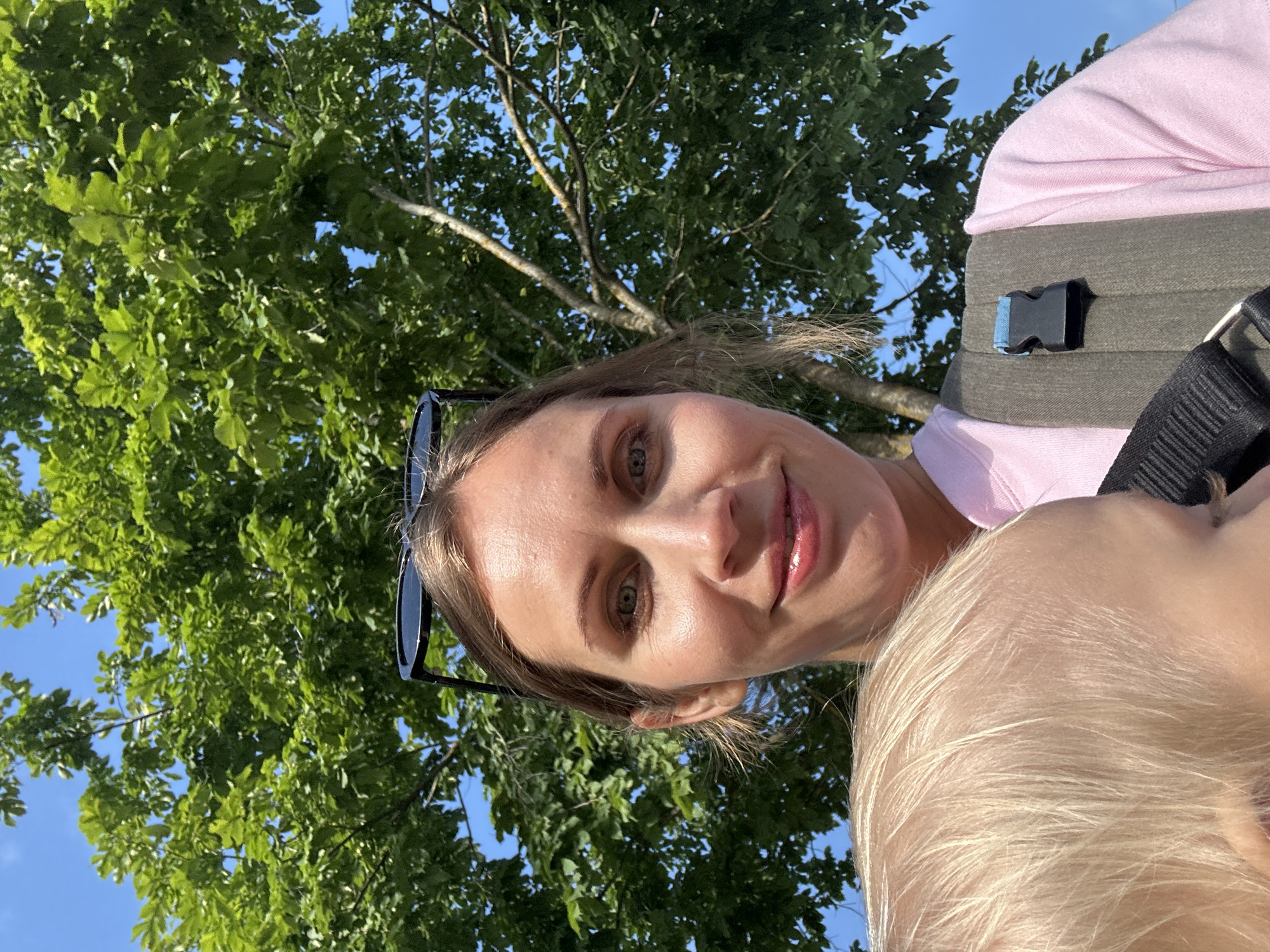 A young woman with blonde hair and green eyes smiles at the camera. She is wearing a pink t-shirt and sunglasses perched on her head. She is holding a baby, whose blonde hair is visible in the foreground. The woman is outdoors, with a large green tree and blue sky behind her. The image was taken in the city of Slantsy, Russia.