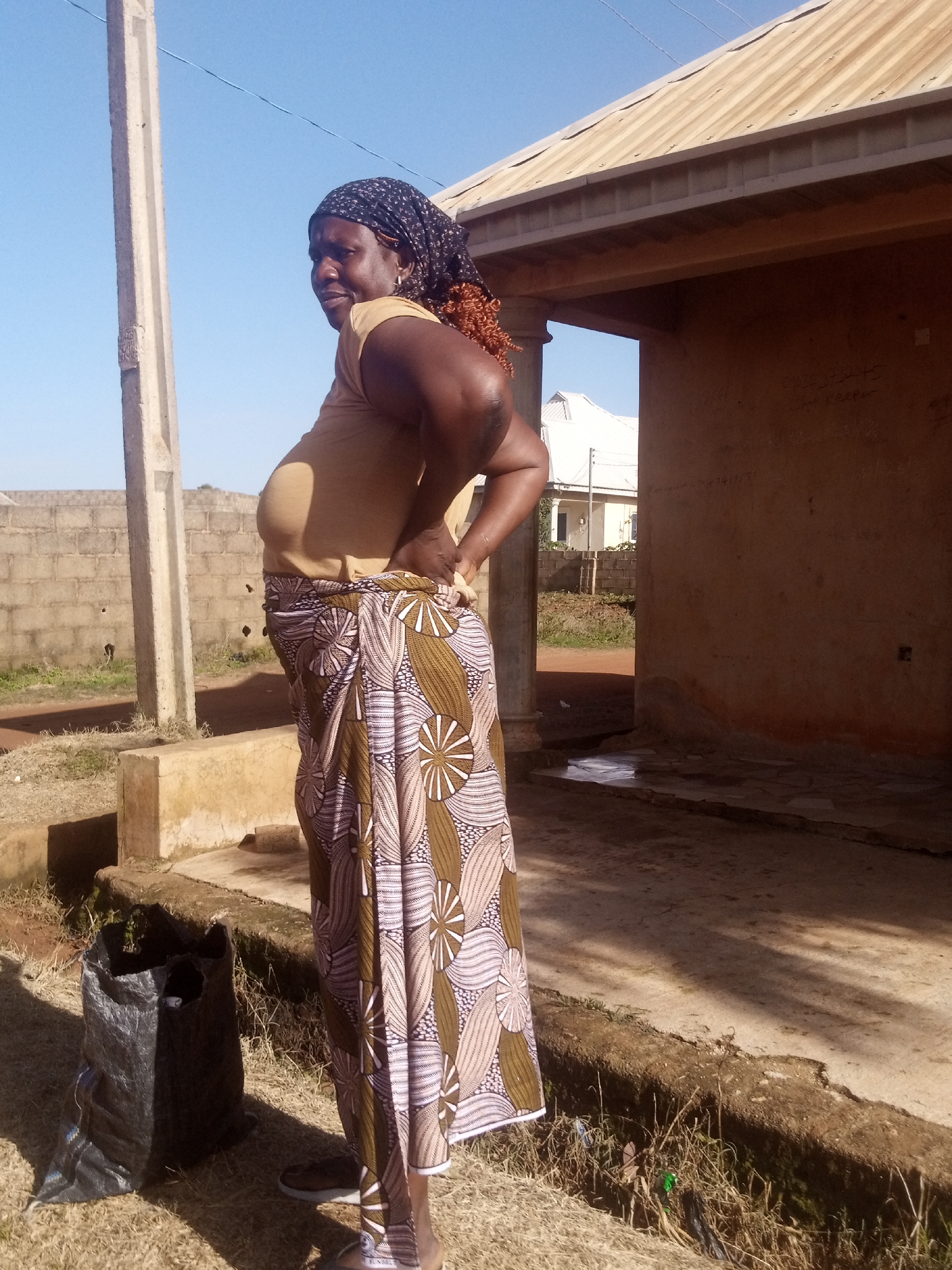 A woman stands in a side profile, facing left, with her left hand on her hip and her right arm bent. She wears a tan t-shirt, a patterned headscarf, and a long, patterned skirt. The woman appears to be of African descent. She has dark curly hair that extends from beneath her headscarf. Her expression is neutral.
The scene is outdoors, with a clear blue sky overhead. To the right of the woman is a building with a tan, corrugated metal roof and light brown walls. The building has an open-fronted structure, creating a shaded area. The ground in front of the building is paved with light-colored tiles. To the left of the woman, there is a concrete pillar and a rough, concrete structure at ground level. Behind the woman is a low brick wall and some dry grass. A black plastic bag lies on the ground to the left of the woman.
The image appears to be taken during the day, as indicated by the bright sunlight and clear sky. The shadows suggest the sun is high in the sky. There is no visible text in the image. The setting seems to be a rural or semi-urban area, possibly in a warm climate.