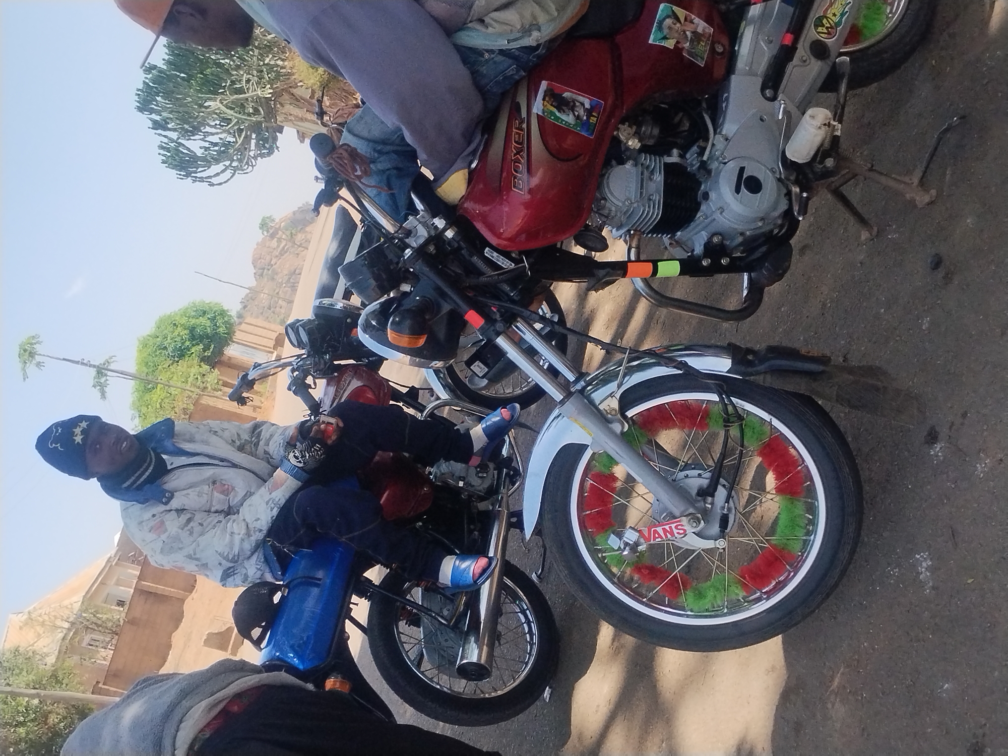 In New Stadium, Nigeria, several motorcycles are parked on a paved surface. One person is sitting on a blue motorcycle with blue and white striped footwear. Another person is sitting on a red "BOXER" branded motorcycle. The front tire of one motorcycle is decorated with red and green material and has a "VANS" logo. Buildings and trees are visible in the background.
