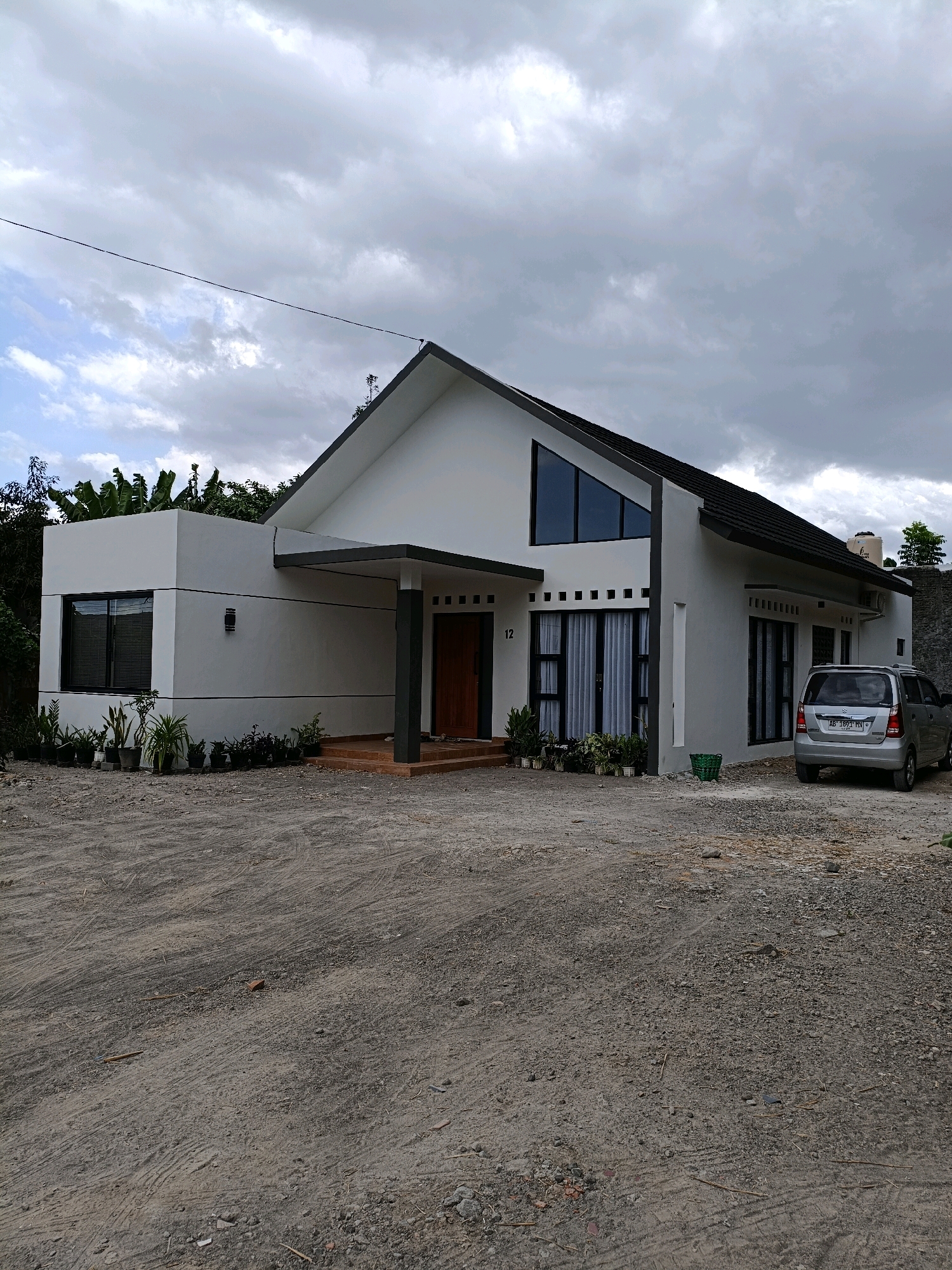 A modern, two-story house with a white exterior and dark gray roof is situated on a gravel lot in Maguwoharjo, Indonesia. The house features large, angular windows, a covered entrance with a wooden door, and several potted plants in front. A silver minivan is parked to the right of the house. The sky is overcast with clouds.