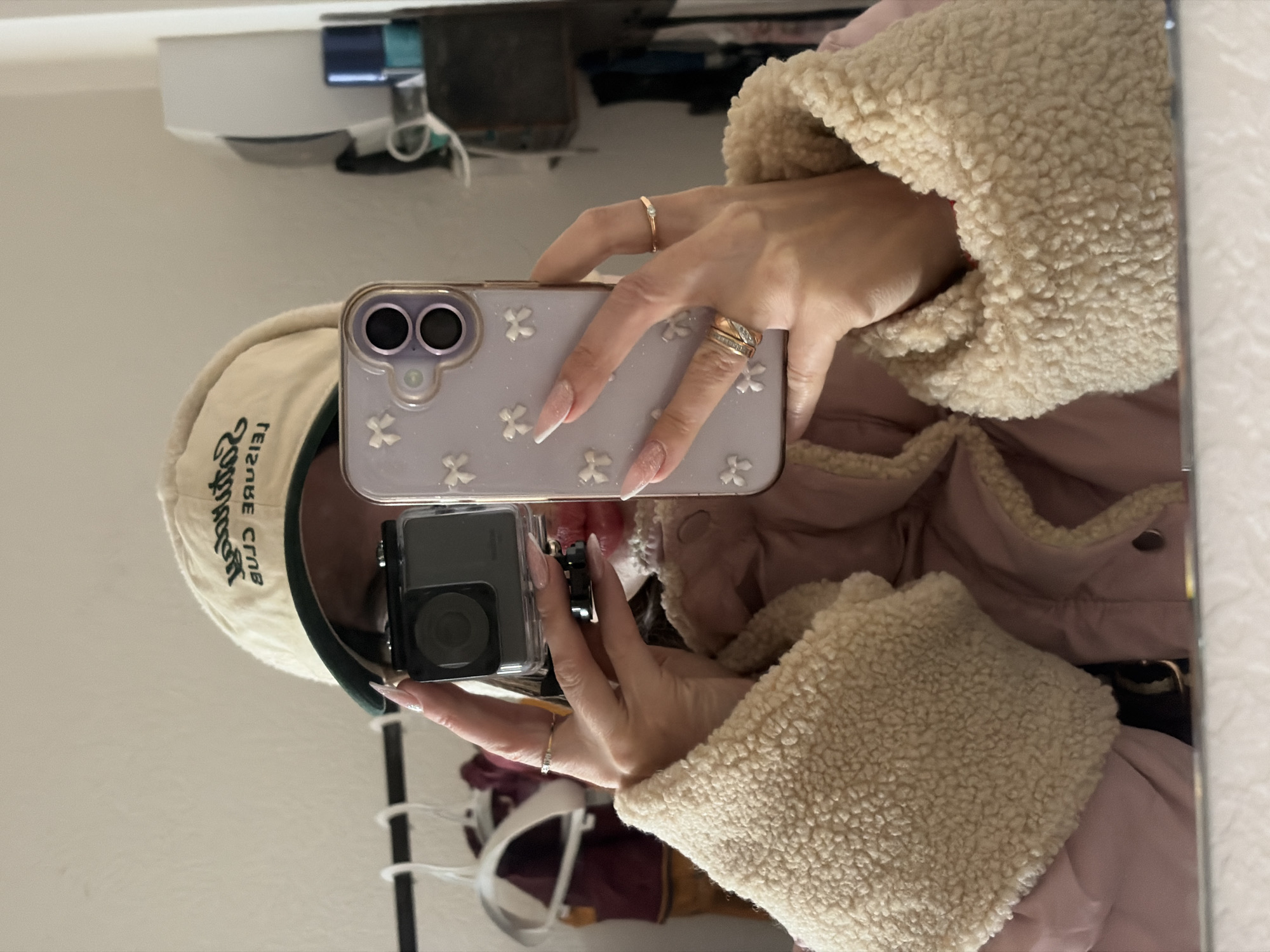 A selfie is being taken in a mirror. The person holding the camera is wearing a tan hat with green writing, a lilac phone case with white flowers, and a coat with cream-colored fleece lining. They are wearing rings on their fingers. The background includes a white wall, a black and white object, and what appears to be clothing on hangers. There is no indication in the image that it was taken in Saint Petersburg, Russia.