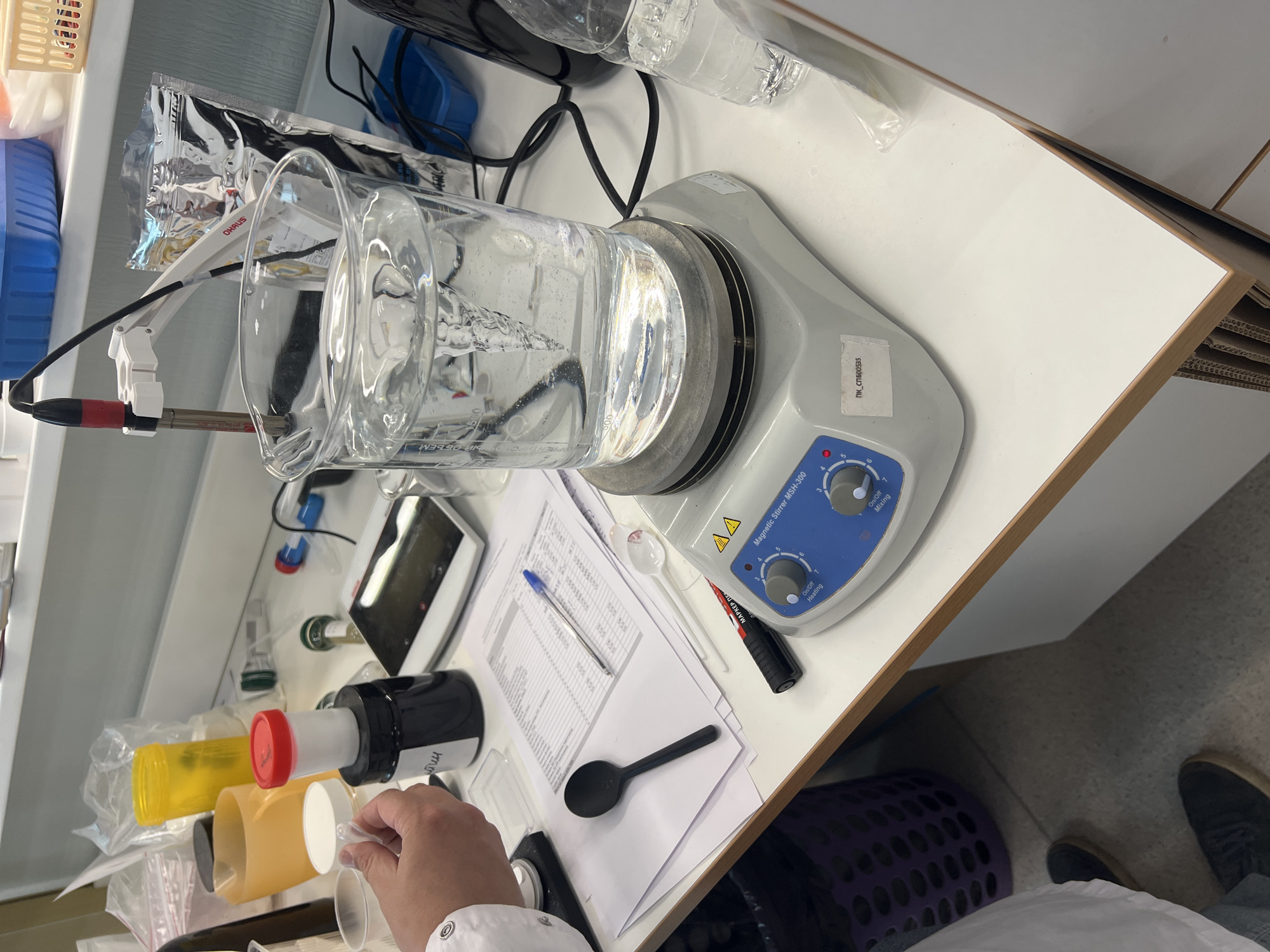 The image shows a laboratory setting. A beaker filled with a clear liquid is sitting on a magnetic stirrer (model MSH-300). An electrode, supported by a white arm with an Ohaus label, is submerged in the liquid. Next to the stirrer are several items, including a pen on a piece of paper filled with data, a black marker, a plastic spoon, and various bottles with colored caps. A person's hand, wearing a lab coat, is holding a small, clear plastic cup, presumably to add something to the beaker. A basket and the person's shoe are visible near the corner of the frame. Location: Novosaratovka, Russia.