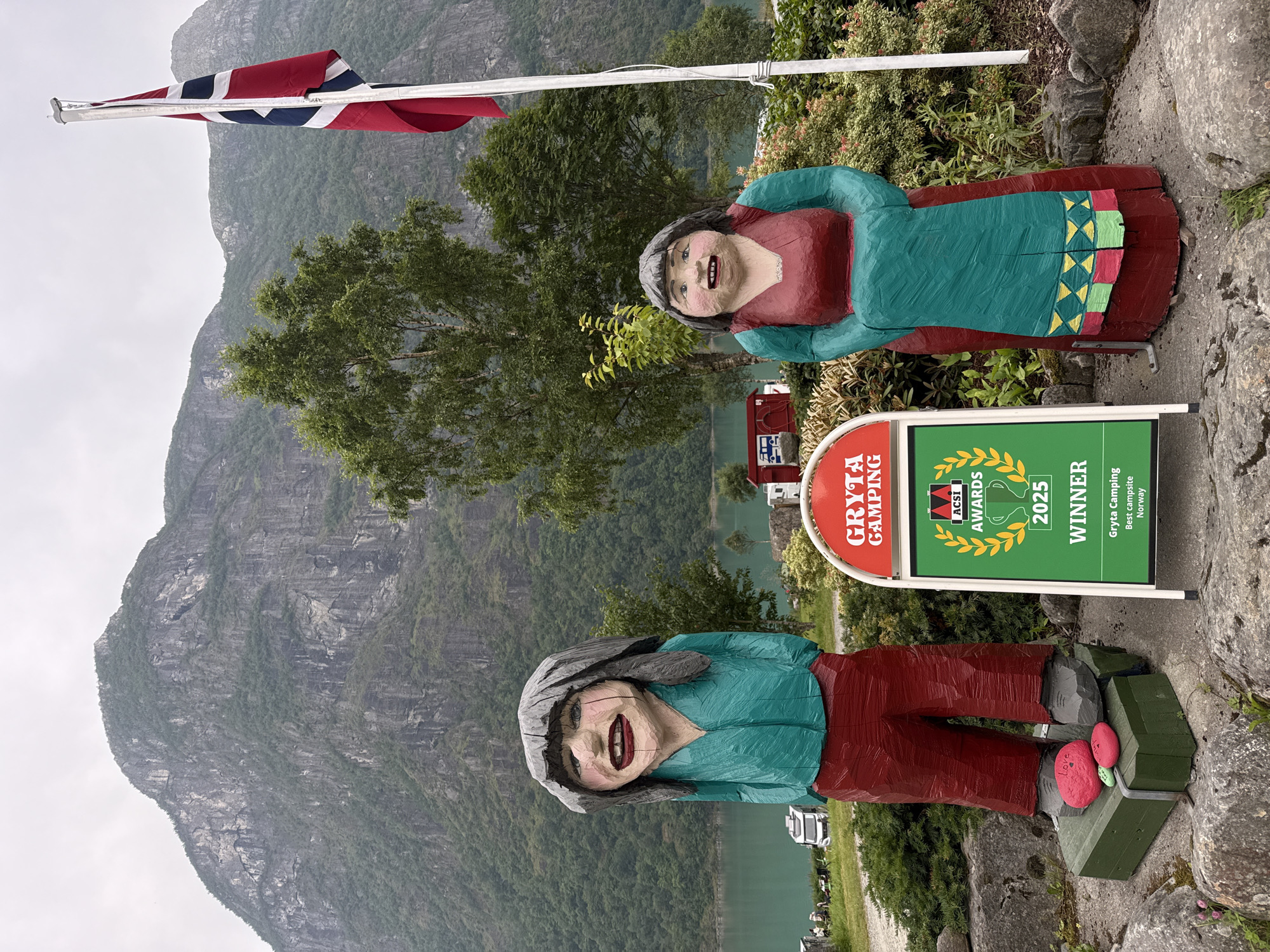 In Olden, Norway, two wooden folk art figures stand outside the Gryta Camping. The figure on the left, dressed in a teal shirt and red pants, has a large smile and grey hair. It stands on a green box with two pink stones, one inscribed with "Love." The figure on the right wears a red dress with teal sleeves and a teal apron adorned with a geometric pattern. Between them, a green and red sign displays "GRYTA CAMPING" and "ACSI AWARDS 2025 WINNER" with "Gryta Camping, Best campsite, Norway" written below. Behind the figures, a calm, turquoise body of water is visible, with a steep, tree-covered mountain range and a cloudy sky in the background. A Norwegian flag flies from a pole to the right of the figures.