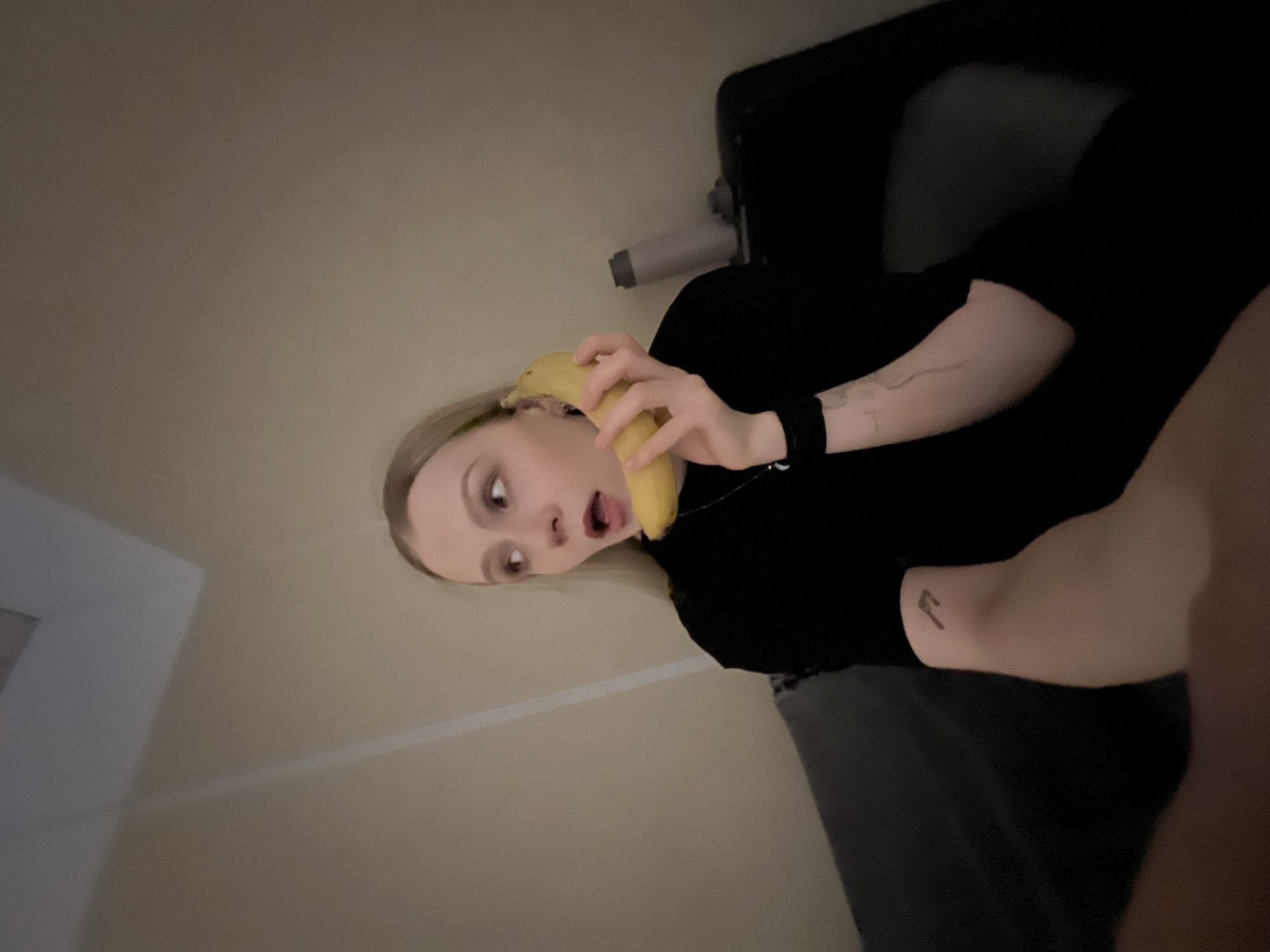 A young, light-skinned woman with blonde hair is holding a banana to her ear, as if using it as a telephone. Her mouth is open in a surprised or shocked expression. She is wearing a black shirt. Visible on her arms are tattoos and a black wristband. She appears to be sitting on a dark gray couch or chair. The background consists of a light-colored wall. No information exists in the provided image to identify Khimki, Russia as the location.
