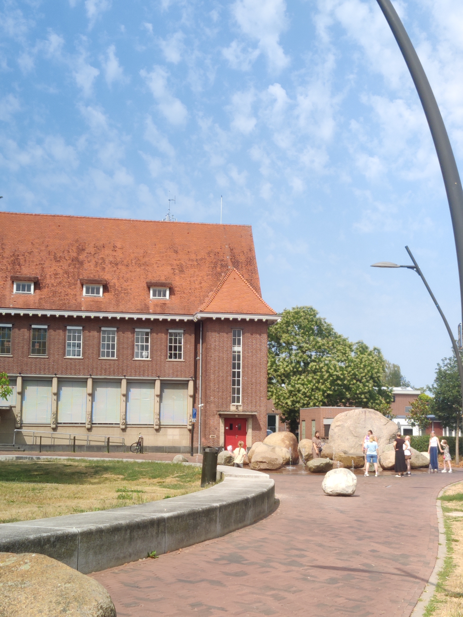 In the city of Winterswijk, Netherlands, the image captures an outdoor scene on a sunny day with a bright blue sky dotted with white clouds. The foreground features a paved area with red brick pavers, leading to a concrete bench. Beyond the bench, a dry, patchy green lawn is visible.
Dominating the left side of the image is a red brick building with a red tiled roof. The building has multiple windows, some with white frames and blinds. Dormer windows are present on the upper level, and a red door is visible on the ground floor.
To the right of the building, a cluster of large, naturalistic rocks forms a water feature, with several people interacting around it. Five young individuals are present: a girl with blonde hair in a white shirt and shorts, a girl in a blue t-shirt and denim shorts, a girl in a white top and dark skirt, a girl in a light-colored top and dark skirt, and a girl in a white top and blue pants. One of the girls is holding a red object, possibly a toy. A dark-colored trash can is situated to the right of the rock formation.
A tall, dark lamppost with a modern design curves over the scene on the far right. In the background, trees with green foliage and other buildings are visible, suggesting an urban or park-like setting. A bicycle is parked near the building on the left.
