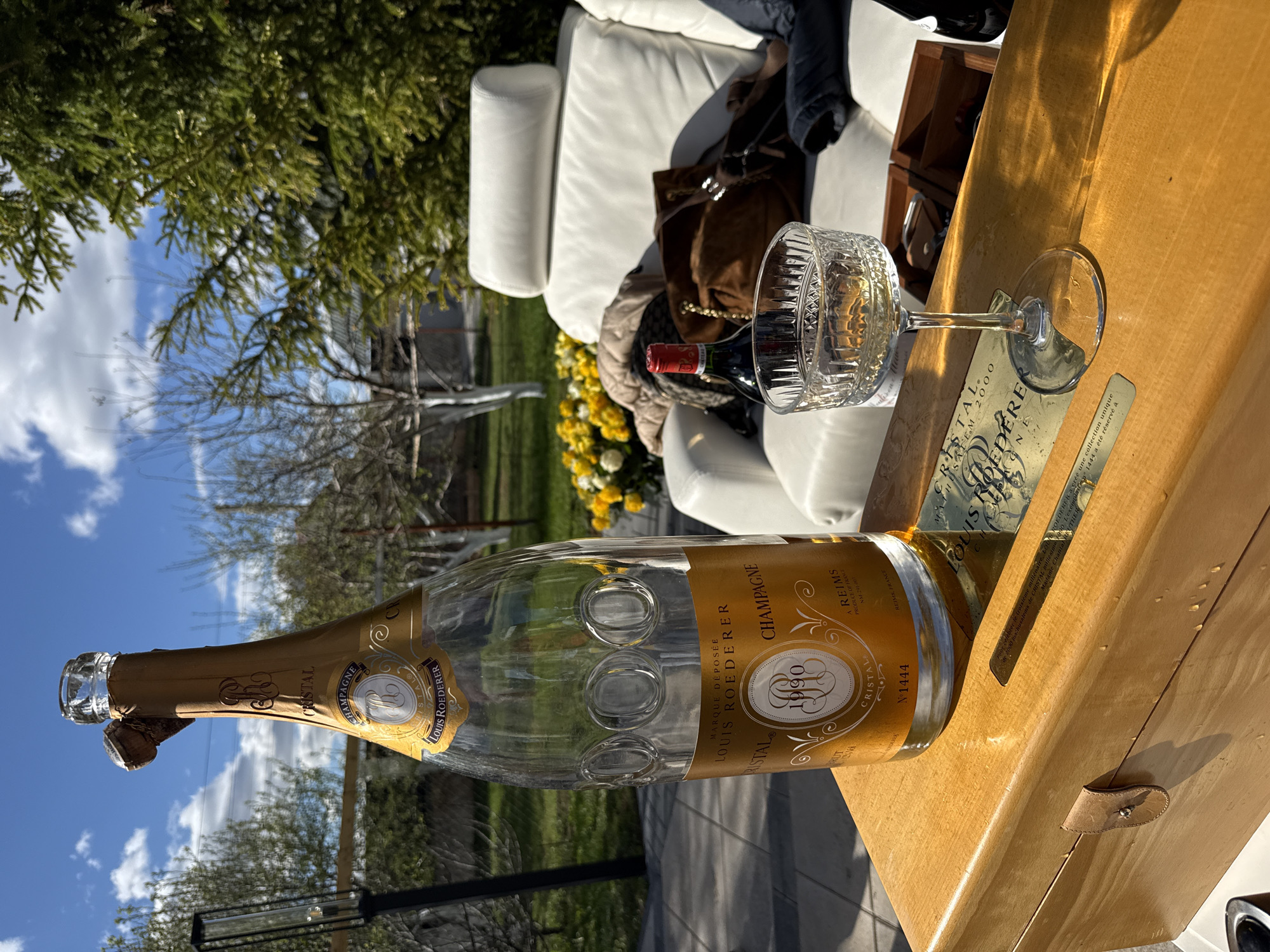 The image shows an outdoor scene on a sunny day, seemingly staged on a patio or deck. An empty bottle of Louis Roederer Cristal champagne (vintage 1990, serial number 1444) lies horizontally on a light-colored wooden surface, possibly a table or a box. Next to the bottle is a crystal glass, appearing to have been used. A metallic card or plaque related to the champagne is also visible on the wooden surface. In the background, a white sofa or chair is situated amidst greenery, including trees and yellow flowers. A purse and an unopened bottle of wine sit on the sofa. The location is unknown.