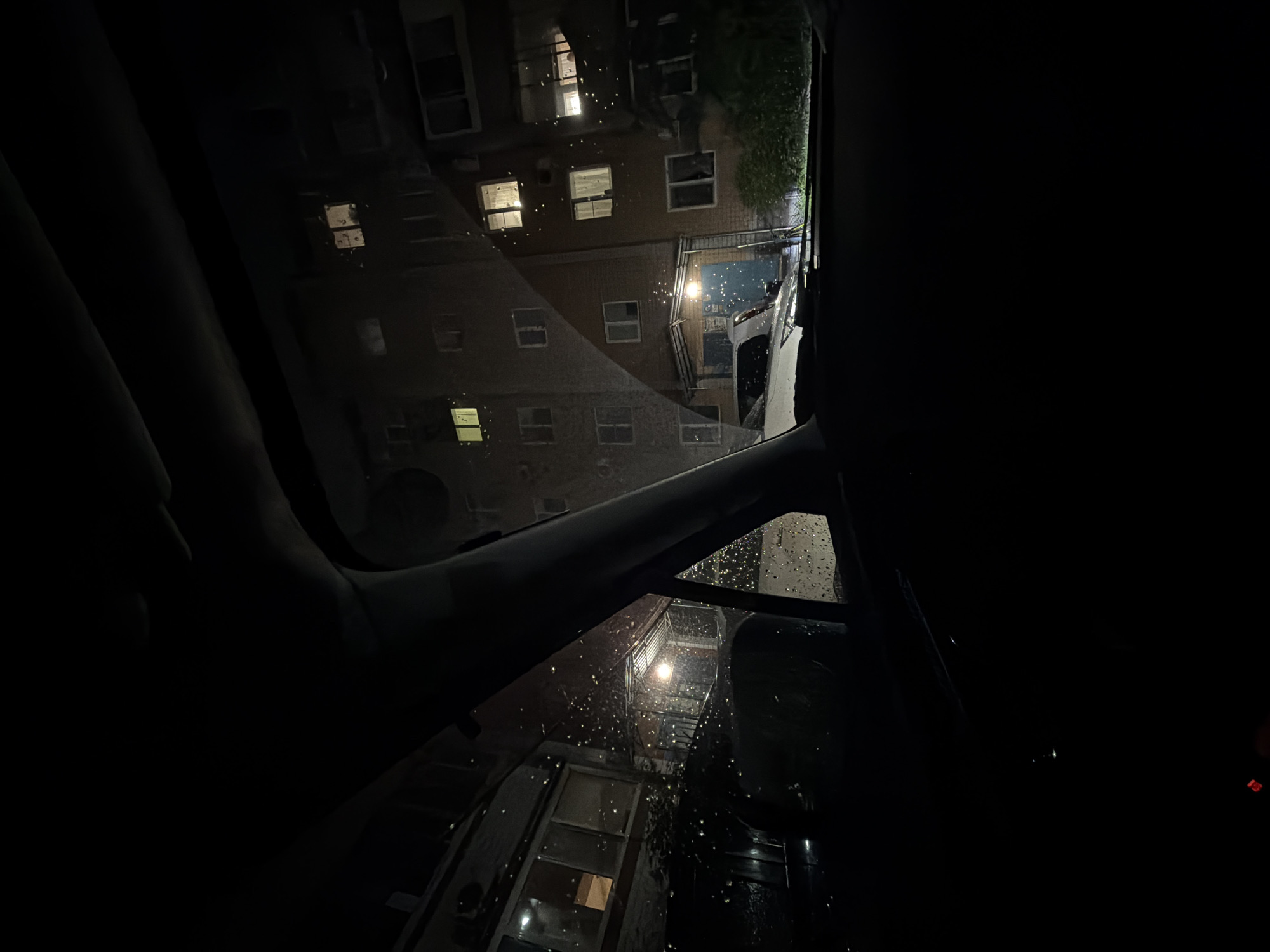 The media file presents a nighttime scene observed from inside a vehicle, likely a car, with a tilted perspective looking up towards a building. The primary subject is a multi-story brick building, possibly an apartment complex, with several windows illuminated from within. An external metal staircase or fire escape structure is visible on the side of the building, and a bright light source, resembling a streetlight, illuminates a portion of the structure and the area beneath the stairs.
The image captures the scene during the night, evidenced by the general darkness and the artificial lights. Numerous small, bright specks are visible on the glass of the vehicle (windshield or sunroof), indicating the presence of rain or water droplets, suggesting a rainy or recently rainy weather condition. Some green foliage is visible along the upper edge of the building, possibly trees or bushes. Elements of the car's interior, such as the roof lining and pillars, frame the view, particularly on the left and right sides, indicating the vantage point is from within the vehicle. No people are visible, and there is no discernible activity taking place. No text is visible in the image. The location context is Birobidzhan, Russia.