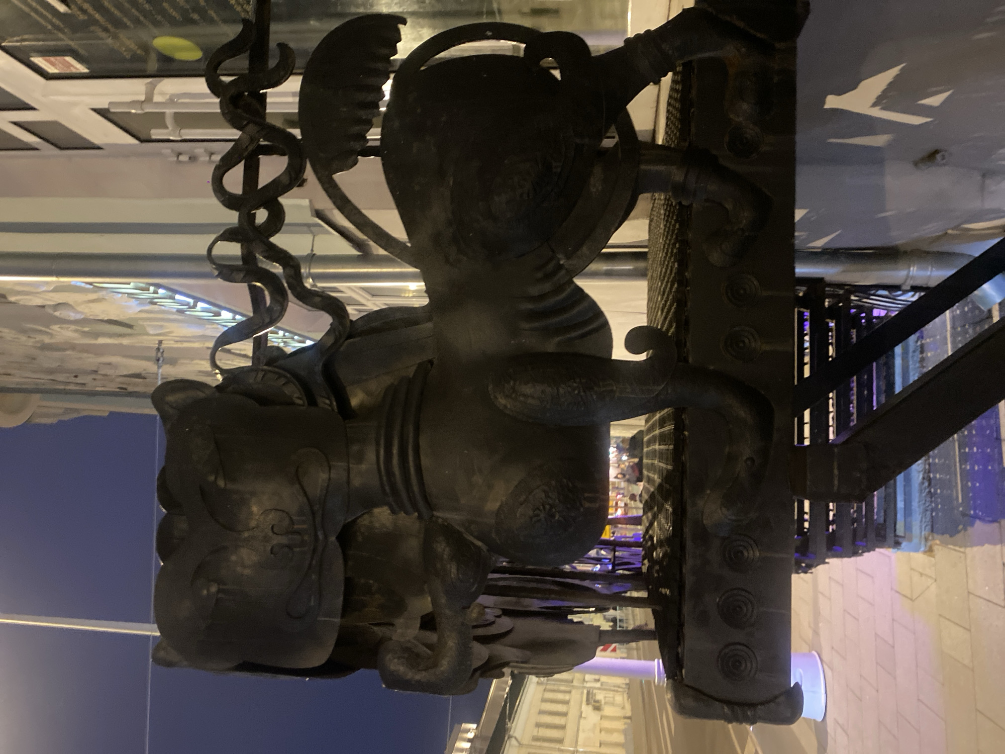The image, rotated 90 degrees clockwise from its natural orientation, presents a detailed view of a dark, ornate metal architectural fixture, likely a decorative element of a lamp post or building facade. The scene is set at night or late dusk in Moscow, Russia, under a dark blue sky, illuminated by artificial lighting.
The primary subject is a robust, dark-colored metal structure, possibly cast iron or bronze, characterized by its intricate design. A central, stylized head of an animal or mythical creature, somewhat resembling a lion or bear, is prominent. This head features curved, ornamental details and is backed by a circular, textured disc. Extending upwards from this central figure is an elegant, coiled element, reminiscent of a serpent or an elaborate decorative scroll.
The lower portion of the structure consists of a sturdy, rectangular metal column adorned with three distinct, raised circular patterns, each featuring a spiral or rosette design. The artificial lighting casts subtle highlights and shadows, emphasizing the texture and three-dimensionality of the metalwork.
In the background, blurred lights and indistinct building shapes suggest a bustling urban environment. Geometric white patterns are visible on an overhead surface, possibly reflections or elements of surrounding architecture. The ground below the fixture is paved, indicating a sidewalk or street. No people are visible, and there is no discernible text in the image. The overall impression is one of historical grandeur and urban artistry.