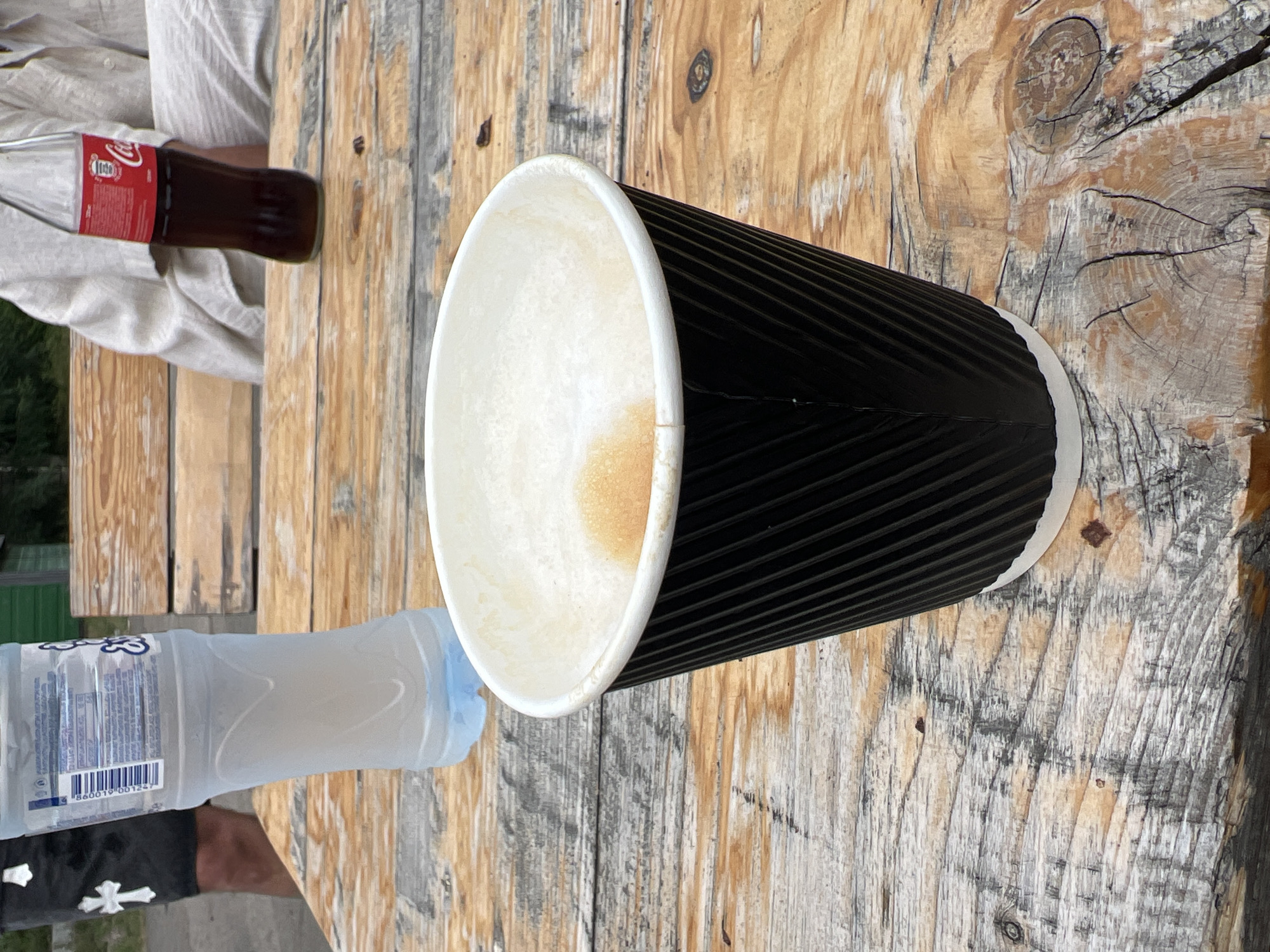A close-up, eye-level shot captures a black paper coffee cup with a white rim, filled with a frothy beverage, resting on a weathered wooden table. The cup is positioned towards the foreground and slightly off-center to the right. Behind it, a partially visible clear plastic bottle of water stands upright, its contents appearing cloudy. To the right of the water bottle, a brown glass bottle of Coca-Cola is also visible, its red label clearly displaying the brand's logo. The background reveals wooden benches, suggesting an outdoor seating area. A person's lower legs and torso are visible on the left, wearing dark shorts with a white cross emblem. Another person, dressed in light-colored clothing, is seated on the right. The overall scene depicts a casual outdoor setting, possibly a cafe or picnic area, in Mukhrovani, Georgia.