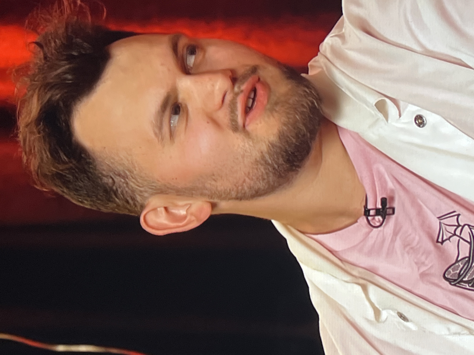 A medium shot shows a man with light brown, messy hair and a short beard. He is looking to his right, with his mouth slightly open as if speaking. He is wearing a pink t-shirt with a graphic design on the front and a white open-front jacket over it. A black microphone clip is attached to the front of his t-shirt. The background is dark, with a red curtain visible behind him. The image appears to be a still from a video recording, possibly an interview or broadcast. The location in Genemuiden, Netherlands, is not discernible from the visual information provided.