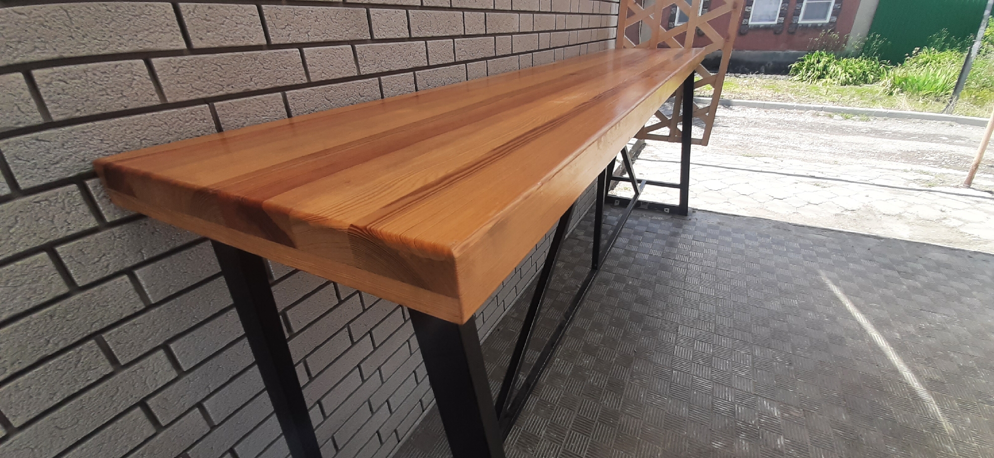The image shows a close-up of a wooden bar top with a black metal frame, positioned against a light gray brick wall. The bar top is made of several wooden planks and has a glossy finish. The legs of the bar are made of black metal and are supported by a patterned rubber floor. In the background, a portion of a wooden chair is visible. Beyond the chair, a street, green vegetation, and a building with windows can be seen under a bright sky. The scene appears to be outdoors, possibly on a patio or balcony. The location is described as Yessentukskaya, Russia, suggesting a specific geographical context.