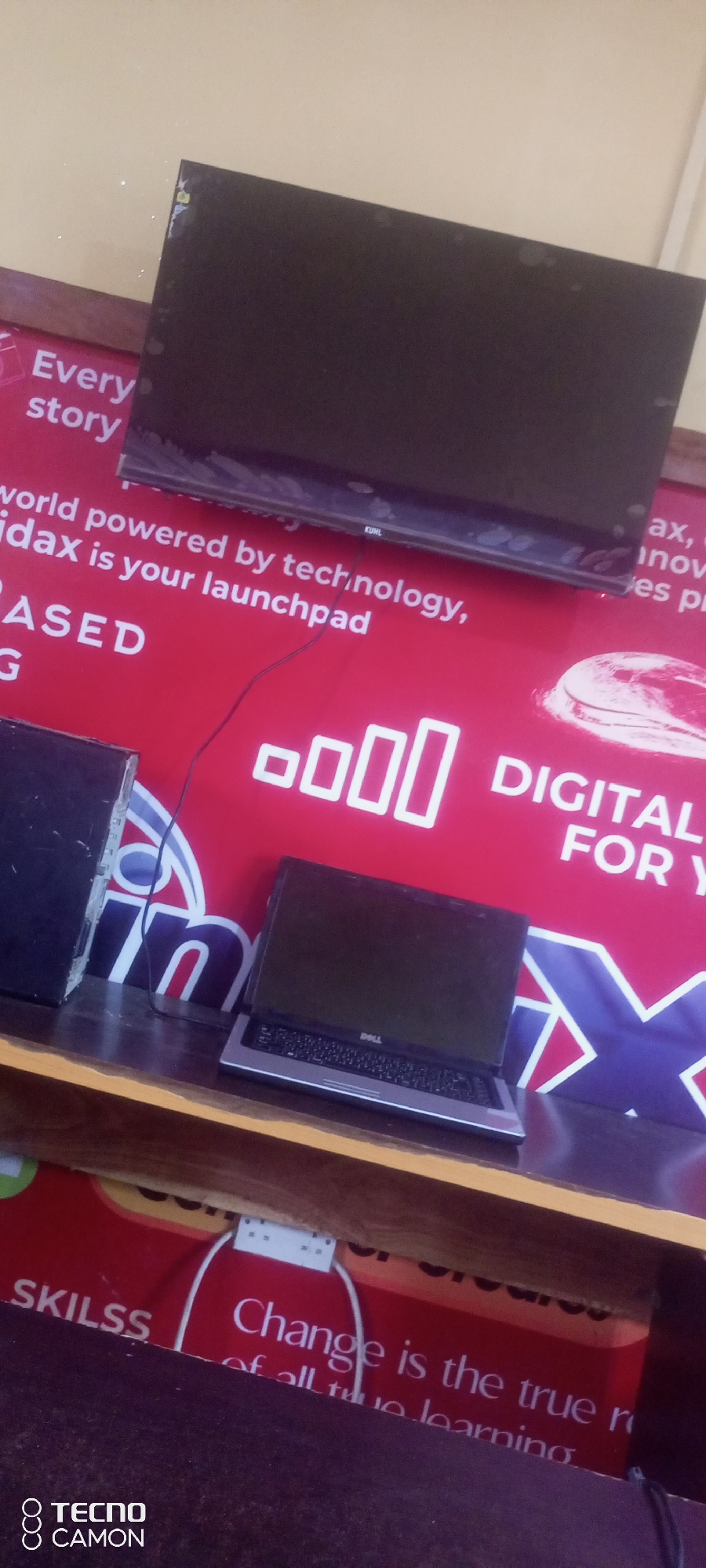 The image shows a display screen mounted on a red backdrop with white text. Below the screen, a Dell laptop is placed on a wooden surface. To the left of the laptop, a black computer tower is visible. The backdrop has several white rectangles arranged in a row, suggesting a signal strength indicator. Further text on the backdrop reads "DIGITAL FOR Y" and "Change is the true r of all true learning". The Tecno Camon logo is present in the bottom left corner of the image. The setting appears to be an indoor commercial space, possibly an electronics store or an office, with the overall impression of a technological or educational theme, potentially in Jalingo, Nigeria, given the mention of Tecno which is popular in the region.