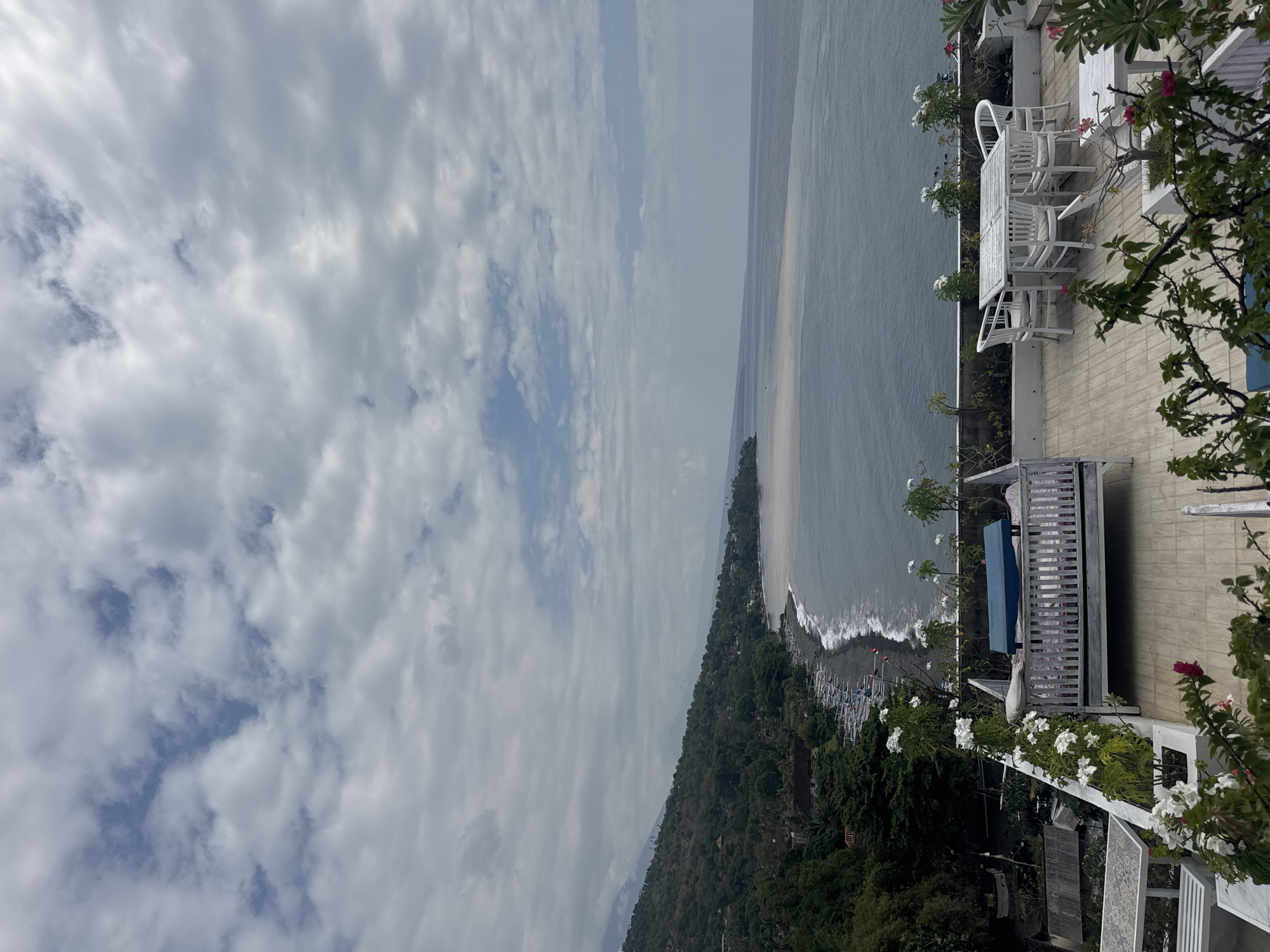 The image shows a view from a rooftop overlooking the ocean in Abang, Indonesia. The rooftop has white outdoor furniture, including a table with four chairs and a daybed with a blue cushion. There are also potted plants with white flowers.
The ocean is visible in the background, with a dark gray color. The coastline is lined with a dark gray, pebbly beach, and a line of small white boats are pulled up on the shore. A lush green hillside with trees and some scattered buildings rises behind the beach. The sky is cloudy with patches of blue visible. The overall atmosphere is peaceful and scenic.