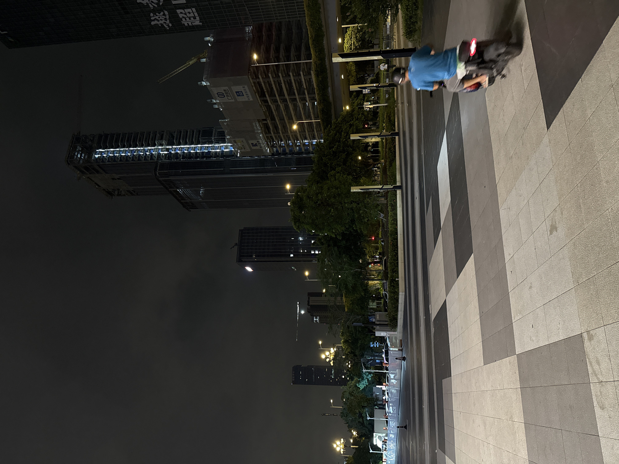 The image depicts an urban night scene in Huangpu District, China, characterized by modern architecture and infrastructure. A person is riding a motorcycle on a patterned pedestrian walkway, moving away from the viewer. The walkway is composed of large, rectangular tiles in varying shades of gray and beige, creating a geometric pattern.
In the background, several tall buildings dominate the skyline. One prominent skyscraper is under construction, with its steel framework visible and some floors illuminated. Other completed skyscrapers are also present, some with lit windows and facades. A yellow construction crane is visible near the building under construction, and another crane is visible in the distance.
Streetlights cast pools of warm light on the walkway and surrounding areas, illuminating trees and bushes. A raised pedestrian bridge or overpass with vertical support columns can be seen spanning across the scene. In the middle ground, a street or road is visible, with some vehicles or temporary barriers suggesting a construction or event zone. The overall atmosphere is one of a bustling city at night, with ongoing development and activity. The sky is dark and overcast, with minimal visible stars.