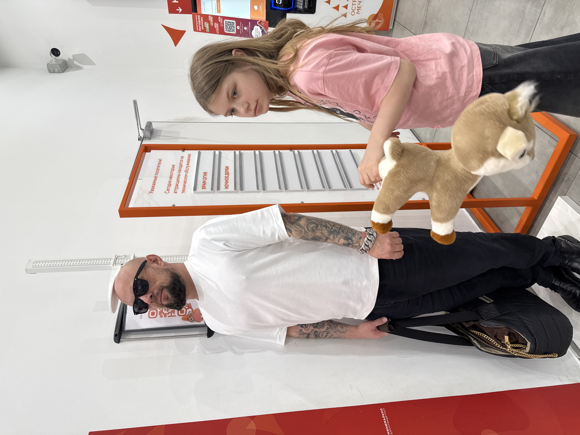 The image shows an indoor scene, likely within an amusement park entrance or information area in Moscow, Russia, given the visible Russian text and context. The setting features bright white walls and a light-colored floor, with orange accents from display stands and floor markings.
In the foreground, a bald man with a beard, wearing black sunglasses, a white T-shirt, black pants, and black shoes, stands. His arms are visibly tattooed, and he wears a thick silver bracelet on his left wrist. He carries a black bag or backpack.
To his right, a young girl with long, light brown hair, dressed in a pink T-shirt and dark pants, leans forward, looking at an orange-framed display stand. She holds a small, light brown and white stuffed deer toy in her hands.
The display stand features several white slots holding signs. A prominent sign on the stand, in Russian, reads: "Уважаемые посетители! Сегодня некоторые аттракционы находятся на техническом обслуживании:" (Dear visitors! Today some attractions are undergoing technical maintenance:). Below this, two specific attractions are listed: "ХРАМ ОГНЯ" (TEMPLE OF FIRE) and "НОЧНОЕ ДЕРБИ" (NIGHT DERBY).
In the background, another sign advertises "БИЛЕТЫ В ПАРК ВЫГОДНЕЕ ОНЛАЙН" (PARK TICKETS CHEAPER ONLINE) and mentions "ОСТРОВ МЕЧТ" (DREAM ISLAND), along with QR codes and pricing information ("от 7000", "до 10000"). A small black security camera is mounted high on the white wall, and a white height measuring stick is also visible. Both the man and the girl appear calm, with the girl seemingly engaged in reading or observing the display.