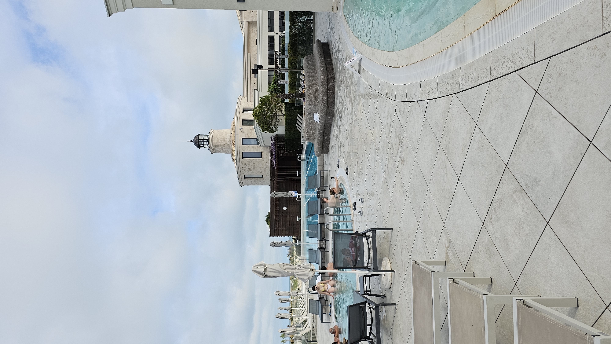 This image depicts a scene at a resort or hotel in Varna, Bulgaria. A multi-level outdoor swimming pool area is present, with several pools of varying sizes and depths. Lounge chairs and tables with umbrellas are placed around the pools. People are swimming and lounging in and around the pools. A lighthouse-like structure stands near the pool area, indicating the resort's aesthetic or theme. The architecture suggests a modern and upscale establishment. The sky is cloudy, suggesting it is daytime but not necessarily sunny. The flooring around the pool is tiled in a geometrical pattern. The image's perspective appears to be taken from an elevated position, possibly from a higher floor of the hotel or resort.