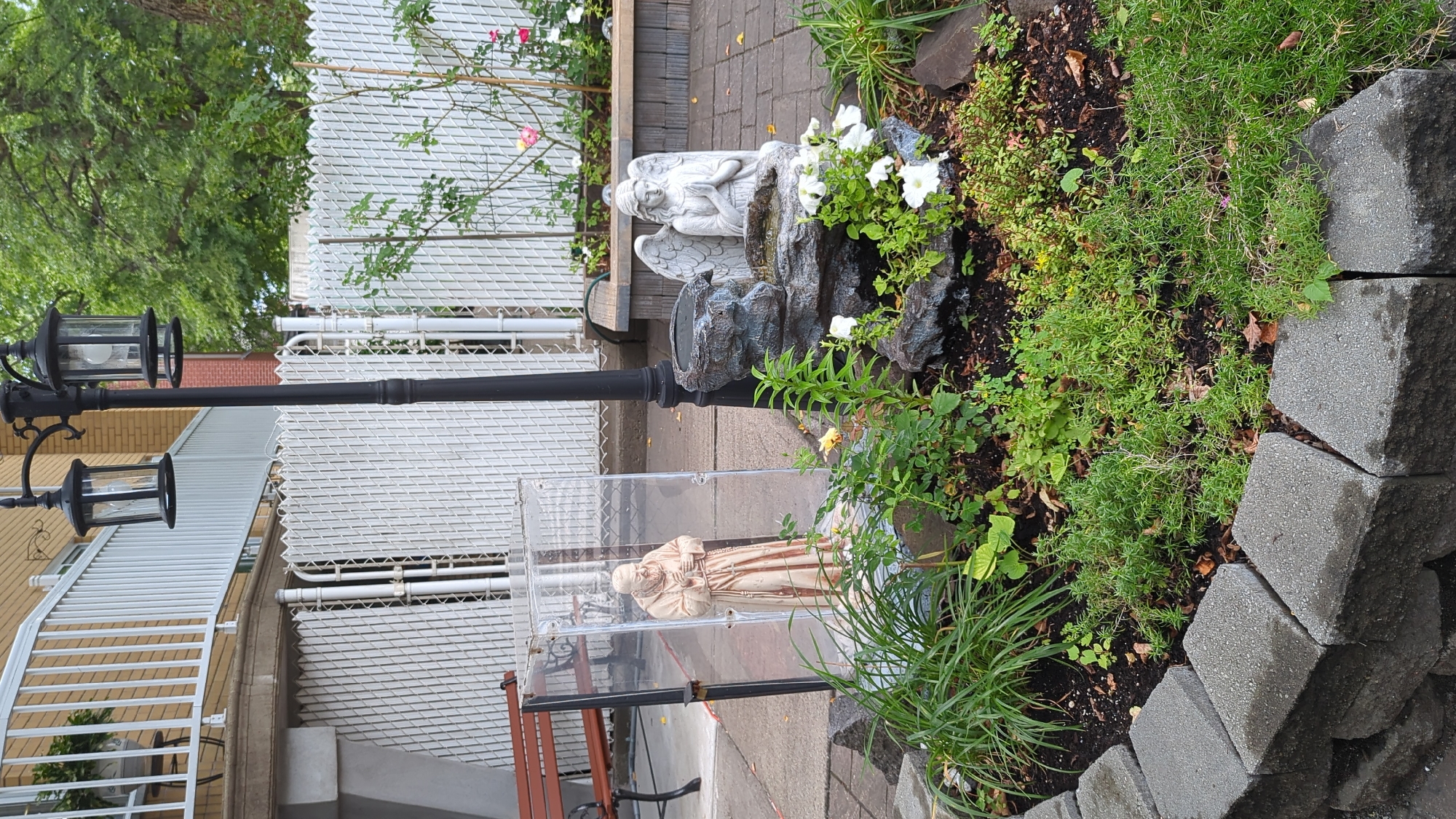 This image depicts an outdoor scene with a religious statue and a small fountain. In the foreground, a statue of a praying monk stands inside a clear protective enclosure. To the right, a small rock fountain features a statue of an angel with folded hands. Both statues are surrounded by lush greenery and small white flowers. A black lamppost with two lanterns stands behind the monk statue. In the background, a white picket fence separates the garden area from a building with a balcony. The ground is paved with grey interlocking stones. The overall impression is a well-maintained garden area, possibly in a residential or public space. The specific location in Montreal, Canada, is not discernible from the image itself.