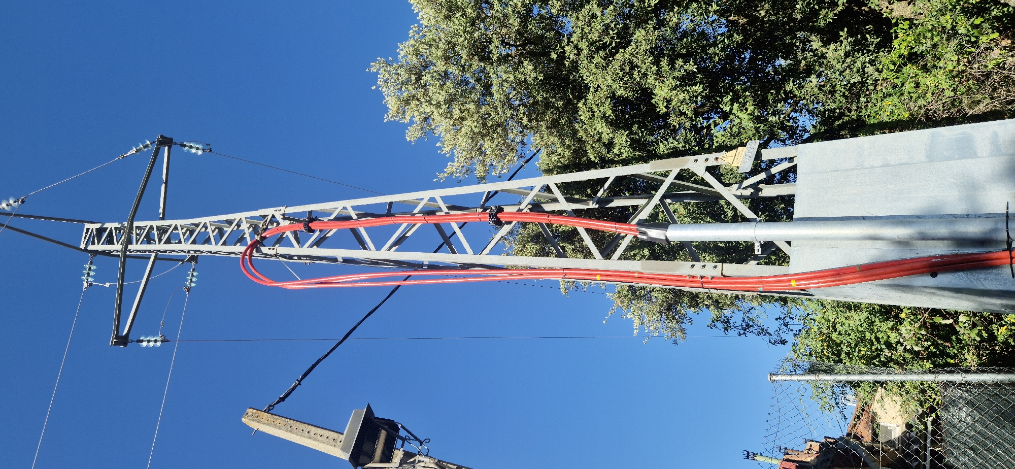 The image shows a close-up, low-angle view of a metal electricity pylon against a clear blue sky. Red conduit pipes are attached vertically to the pylon with black clamps. The pylon has insulators and wires extending from its top. To the left, a concrete support structure with a metallic box is visible, connected to the pylon by a black wire. In the background, lush green trees with dense foliage are present. A chain-link fence and some vegetation are in the lower left foreground. The scene appears to be outdoors. A yellow warning sign with black text is affixed to the pylon. The location is identified as Navaluenga, Spain.