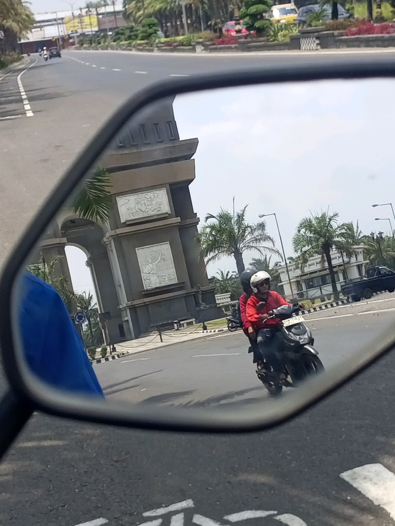 The image is a reflection in a motorcycle's side mirror, showing a view of the city of Ngasem, Indonesia. In the foreground of the reflection, a motorcycle with two occupants is visible, with the rider wearing a red shirt and white helmet, and the passenger wearing a black helmet. The motorcycle is traveling on a paved road. Behind the motorcycle, a large, ornate archway with relief carvings is prominent. Palm trees and other tropical vegetation line the street, with buildings and streetlights also present in the background. The overall scene depicts a typical urban streetscape in a tropical climate.