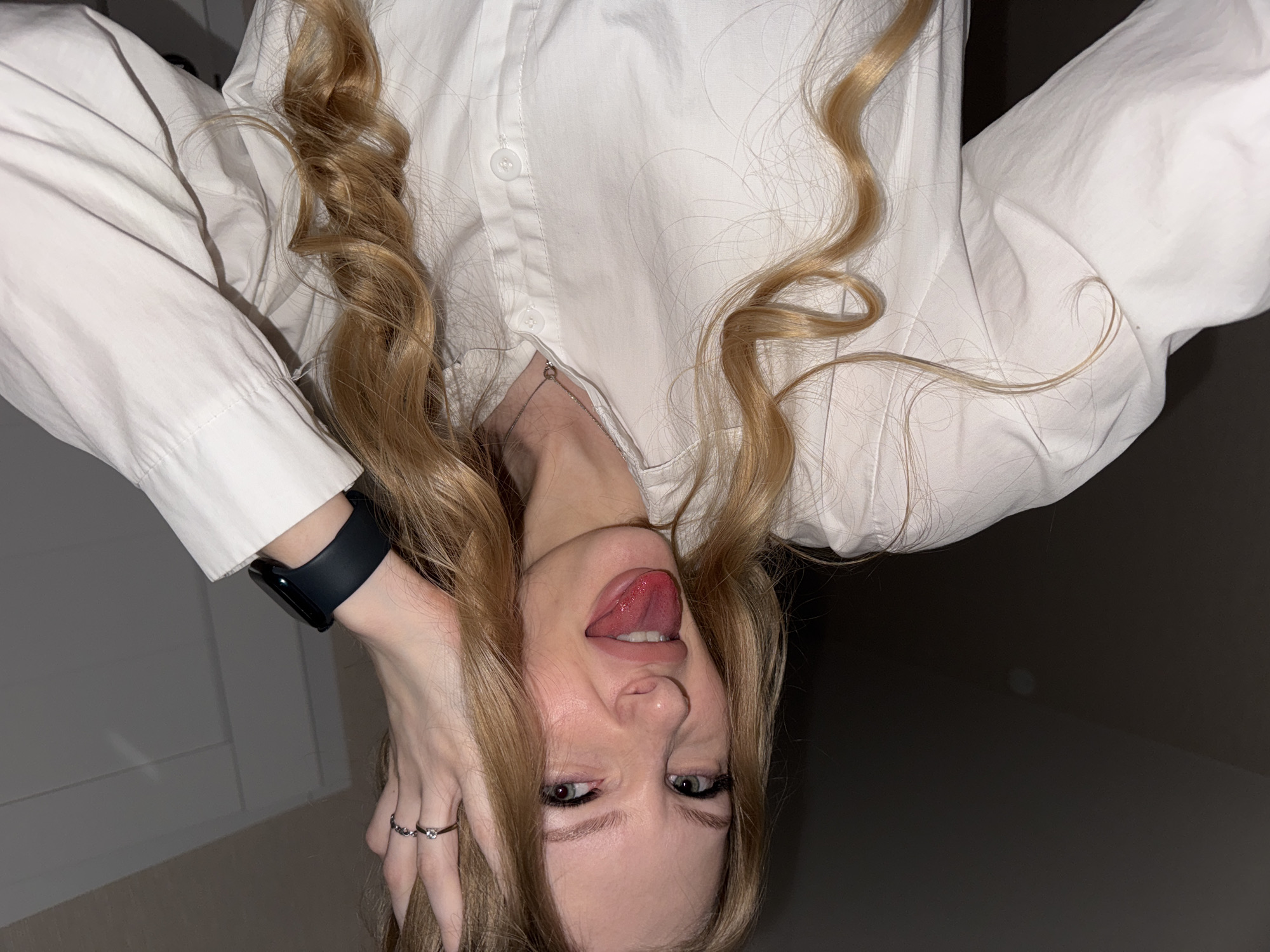 The image shows a young woman with blonde hair wearing a white button-up shirt and a black smartwatch. She is sticking her tongue out and touching her hair with her left hand. There are two rings on her left hand. The image is a close-up, and the background is dark. There is no indication of the city of Perm, Russia.