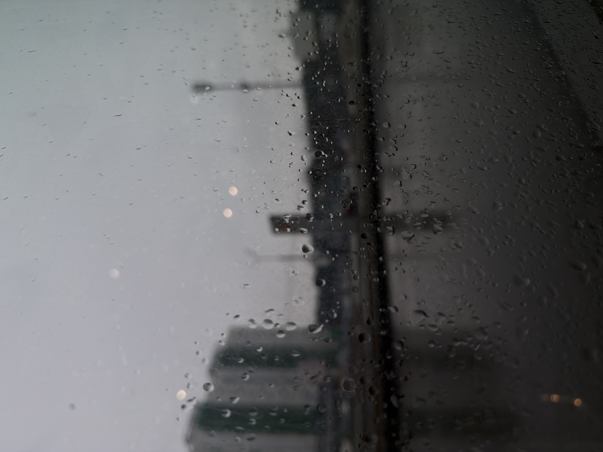 The image shows a rain-streaked window with a blurred cityscape visible through the raindrops. On the left side, the scene is lighter and more overcast, with indistinct shapes suggesting buildings and streetlights. The right side of the window is darker, with denser condensation obscuring the view. The cityscape appears to be blurred and out of focus, indicating it is viewed through the wet glass. The location is unspecified and the presence of people, interactions, or specific objects from Izhevsk, Russia cannot be confirmed.