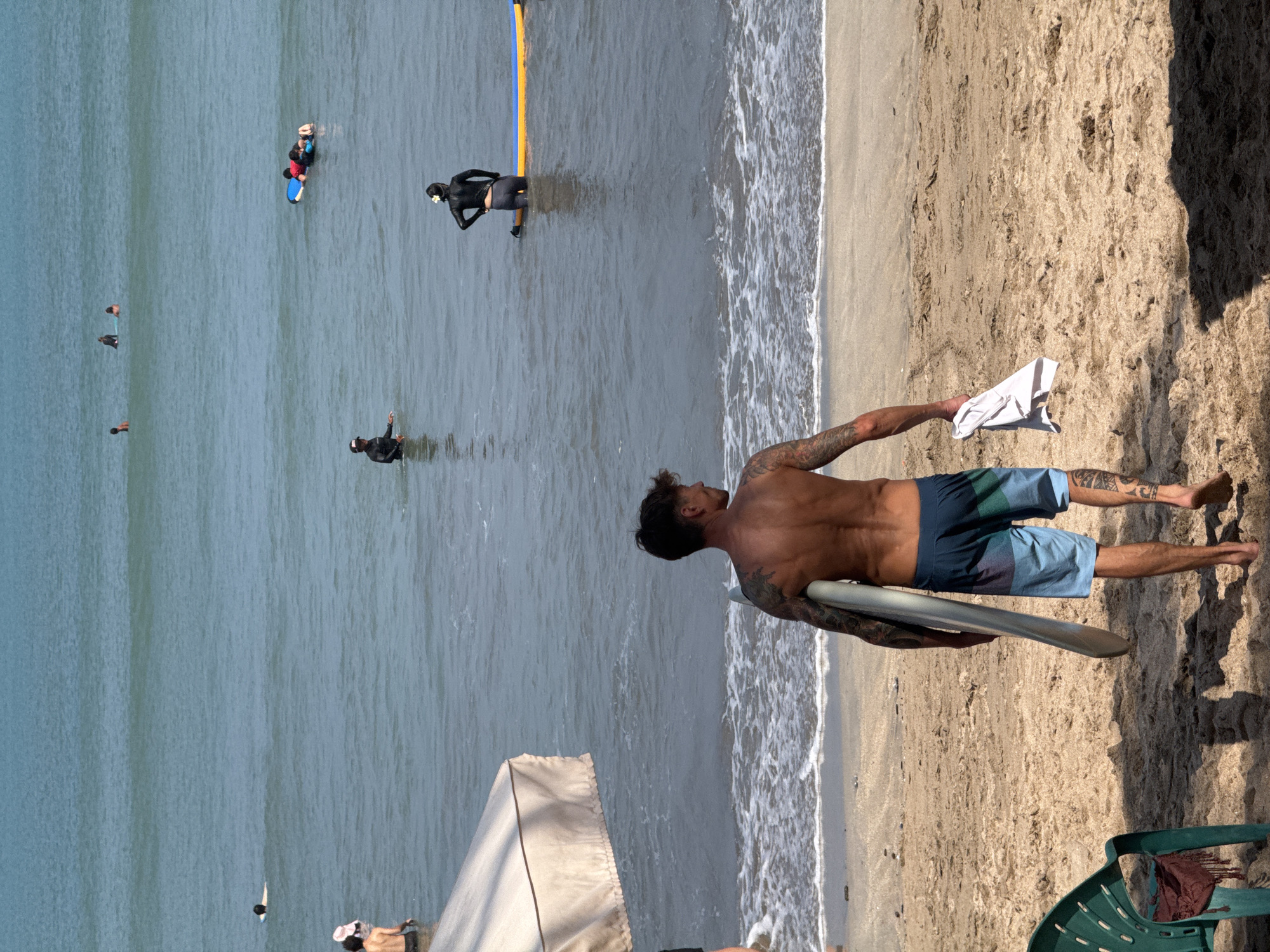 An overhead shot looks down on a beach in Kuta, Indonesia, on a bright, sunny day. A man, his back to the camera, walks from the shore into the ocean, holding a surfboard and a white towel. He is wearing multi-colored swim trunks and has numerous tattoos on his arms and legs. Several other people are in the water, some on surfboards, in the distance. A beach umbrella is visible to the left of the frame. The water is calm with small waves.