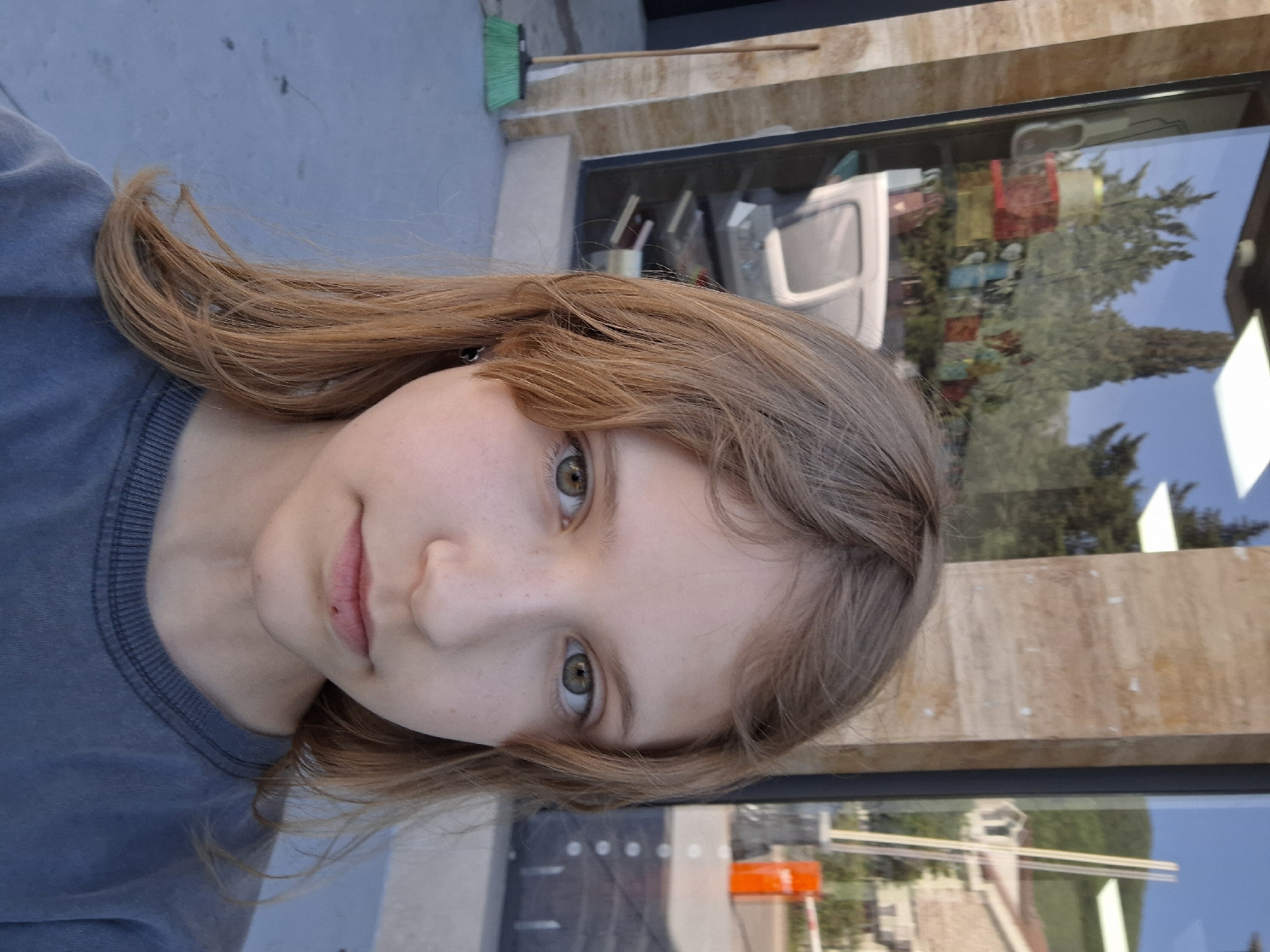 A young girl with light brown hair and green eyes looks directly at the camera, positioned slightly off-center. She wears a gray t-shirt. Behind her is a commercial building with a large window reflecting trees and buildings, suggesting an urban environment. A green broom rests on the building's exterior. The reflected scenery and architecture suggest that the location might be in an urbanized area of Budva, Montenegro.